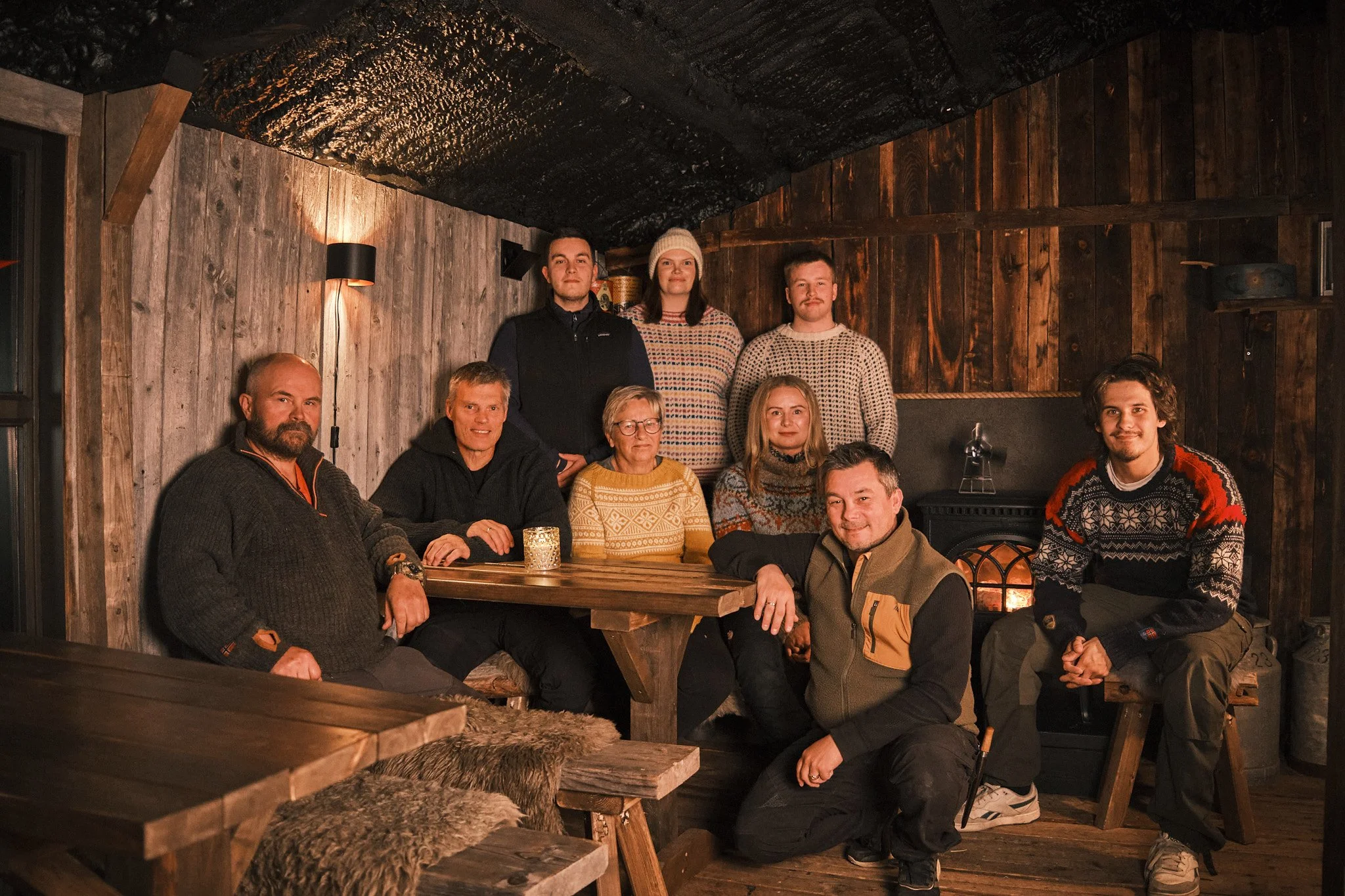 Group of nine people gathered in a cozy, rustic room with wooden walls and ceiling, sitting and standing around a wooden table near a fireplace, dressed in warm sweaters and jackets.