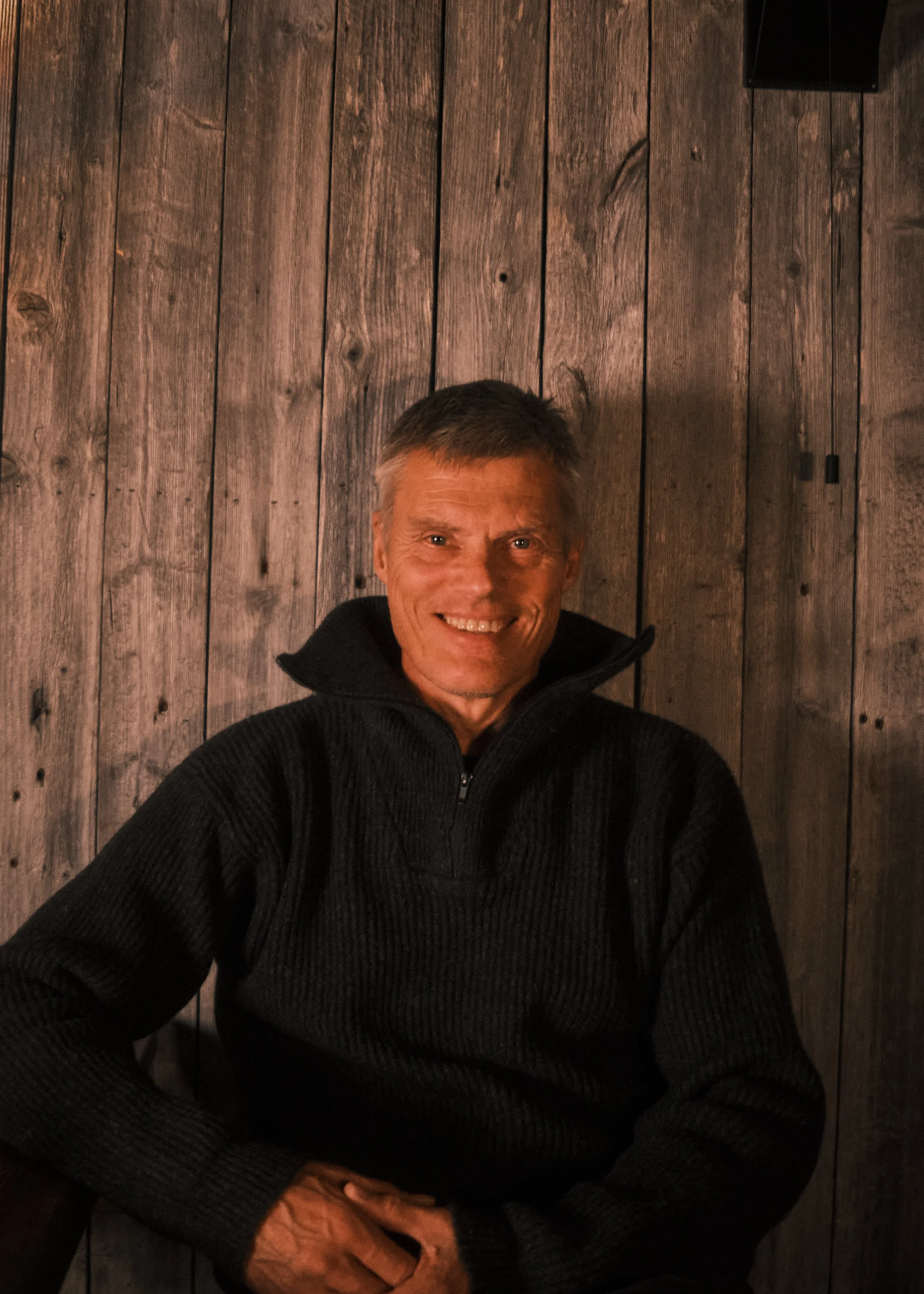 A smiling middle-aged man with short gray hair, wearing a black fleece pullover, sitting in front of a wooden wall.