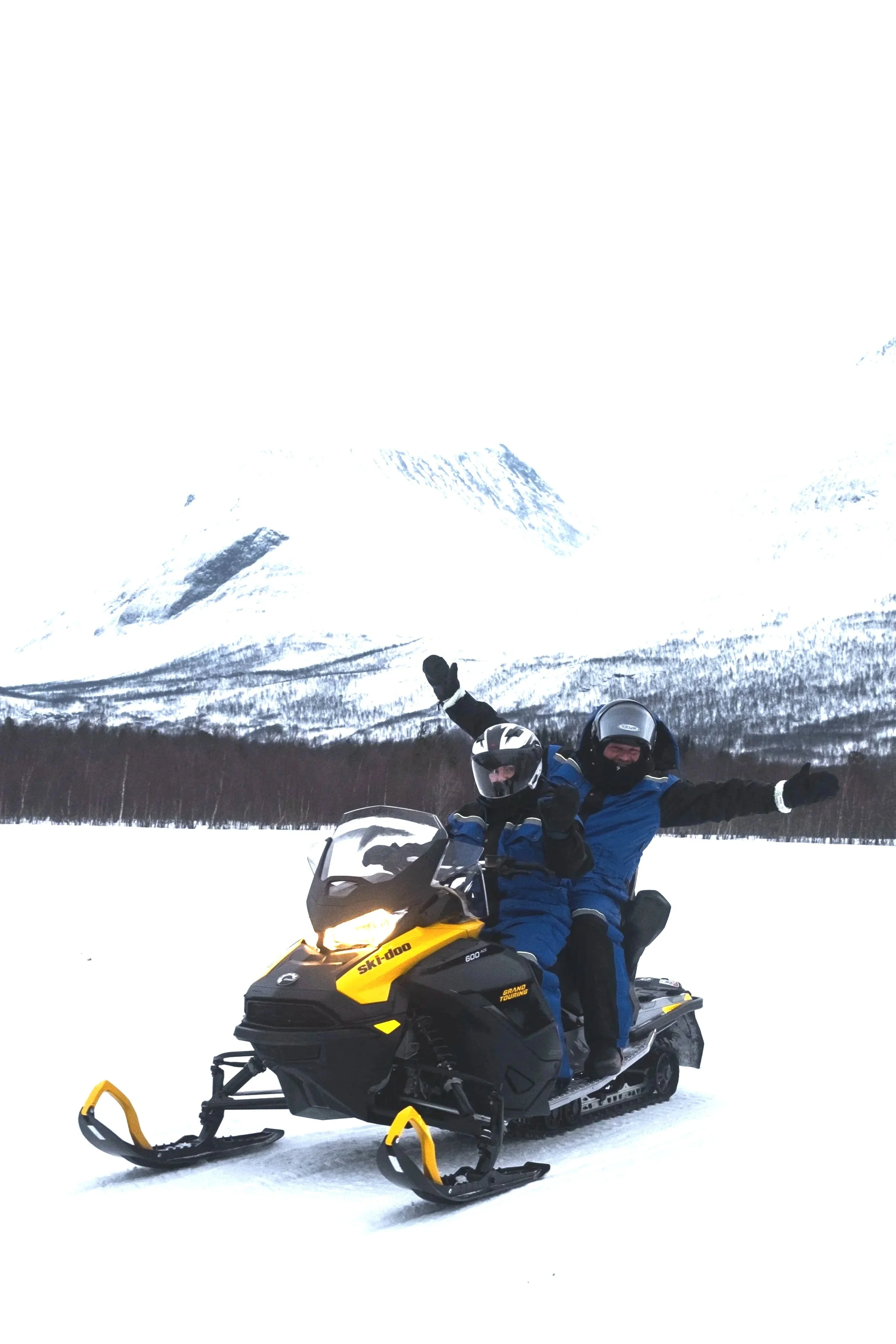 Two people riding a yellow and black Ski-Doo snowmobile on a snowy landscape with a mountain in the background. Both are wearing helmets and winter gear, with one person giving a fist pump and the other raising an arm in celebration.