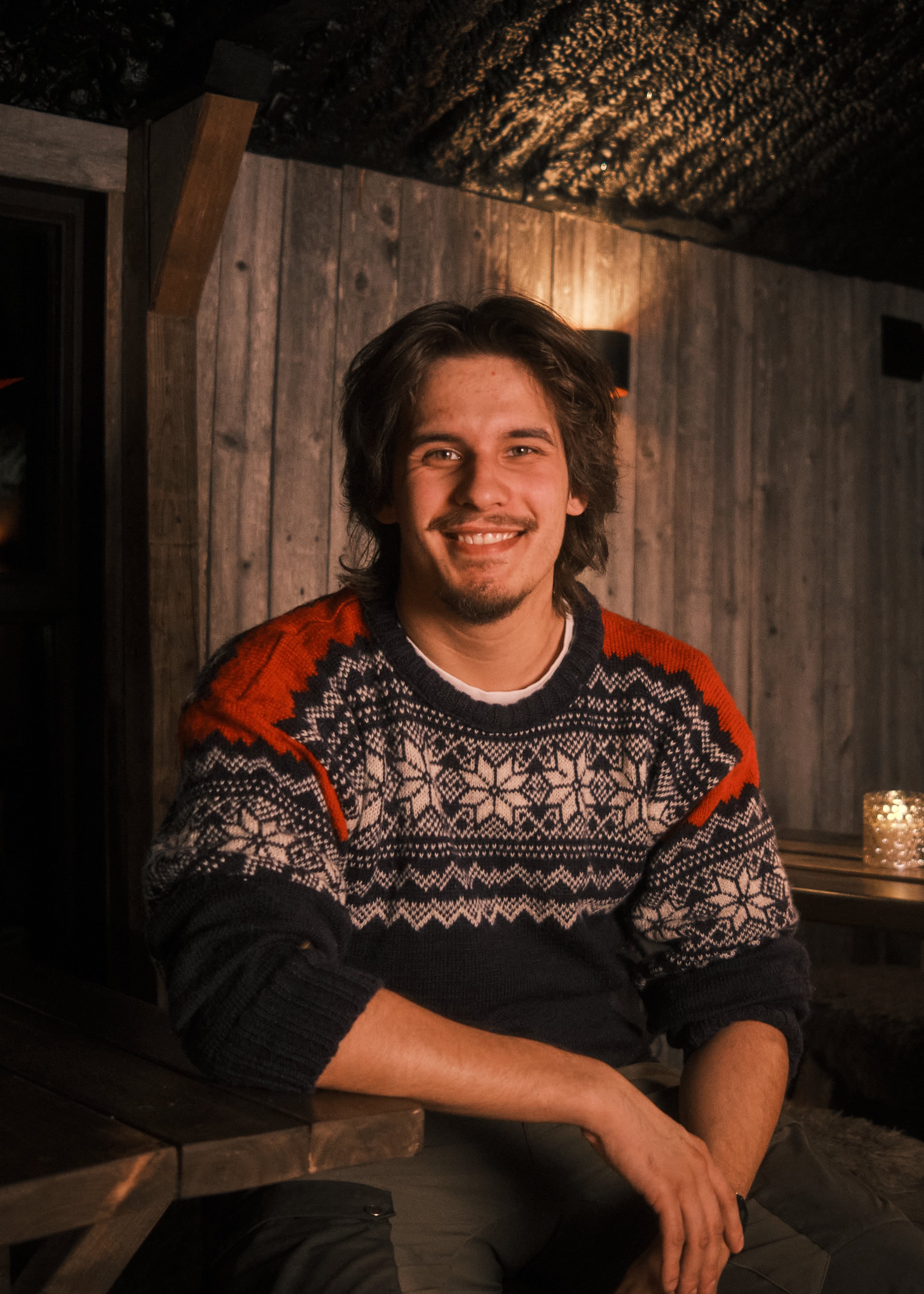 A young man with long brown hair, smiling, sitting at a wooden table in a cozy, wood-paneled room with warm lighting.