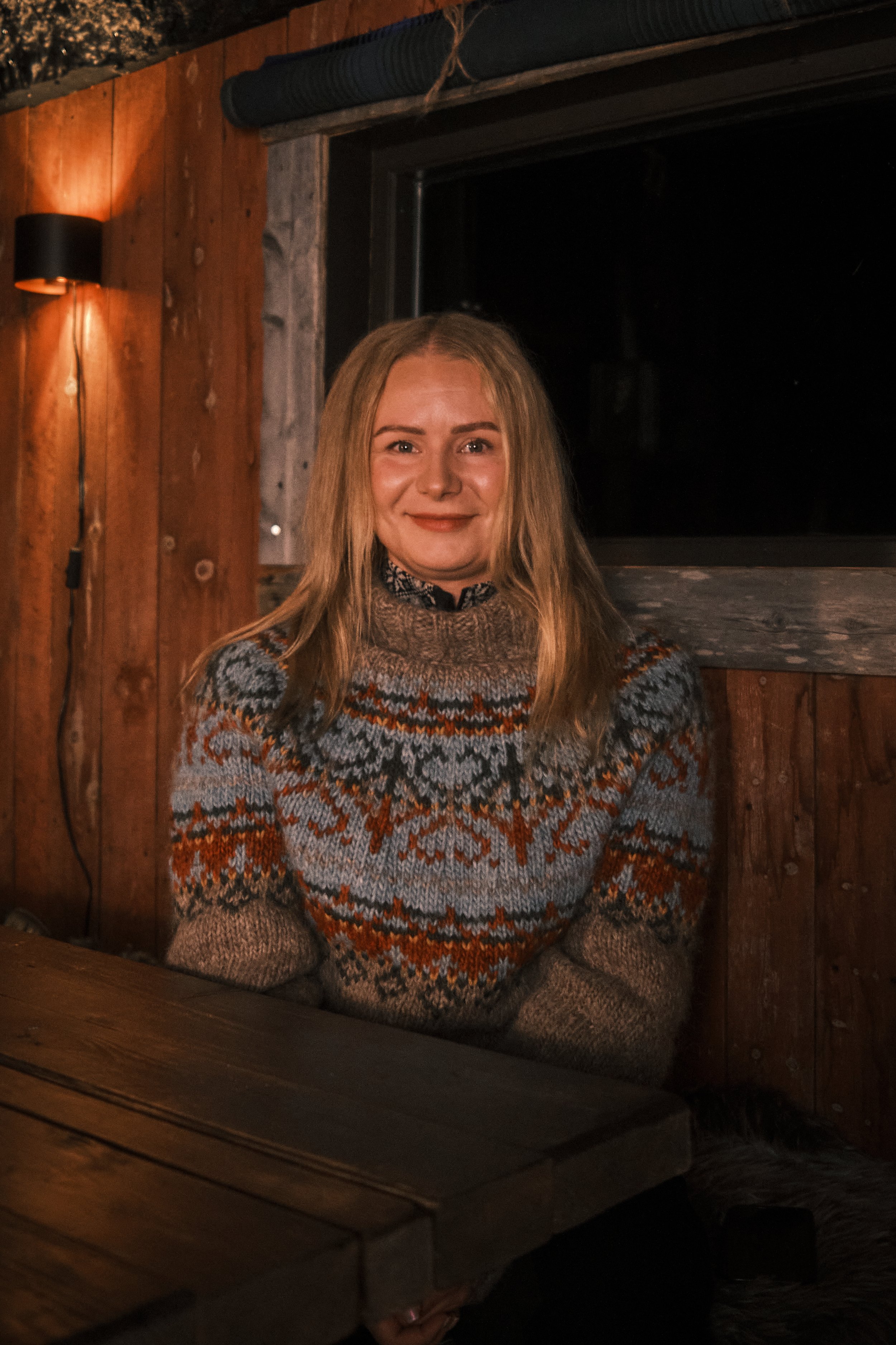 A woman sitting at a wooden table inside a cozy cabin, wearing a colorful patterned sweater and smiling.