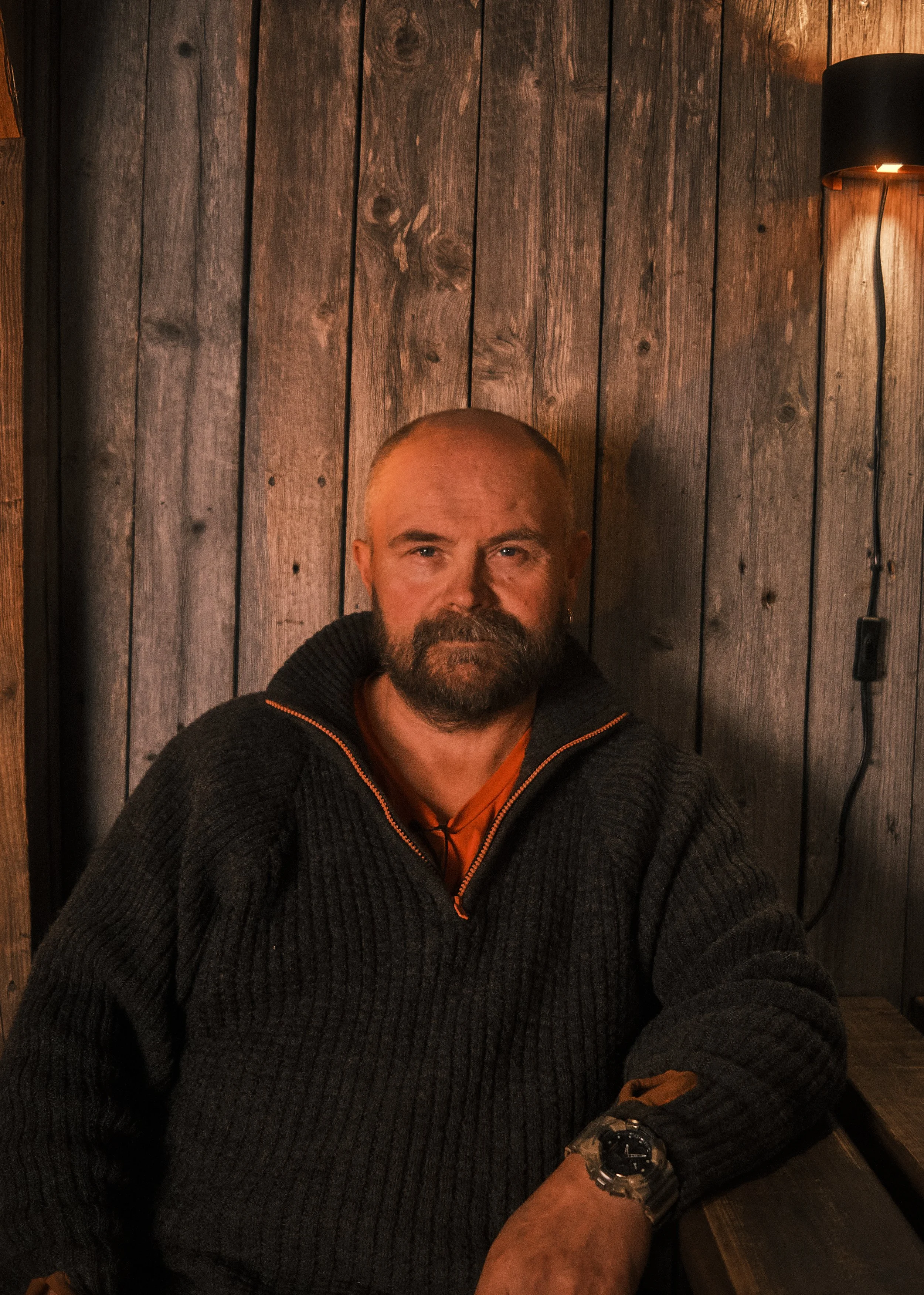 A man with a beard and absent hair, wearing a dark knit sweater with a partially unzipped collar, sitting against a wooden wall in a dimly lit room.