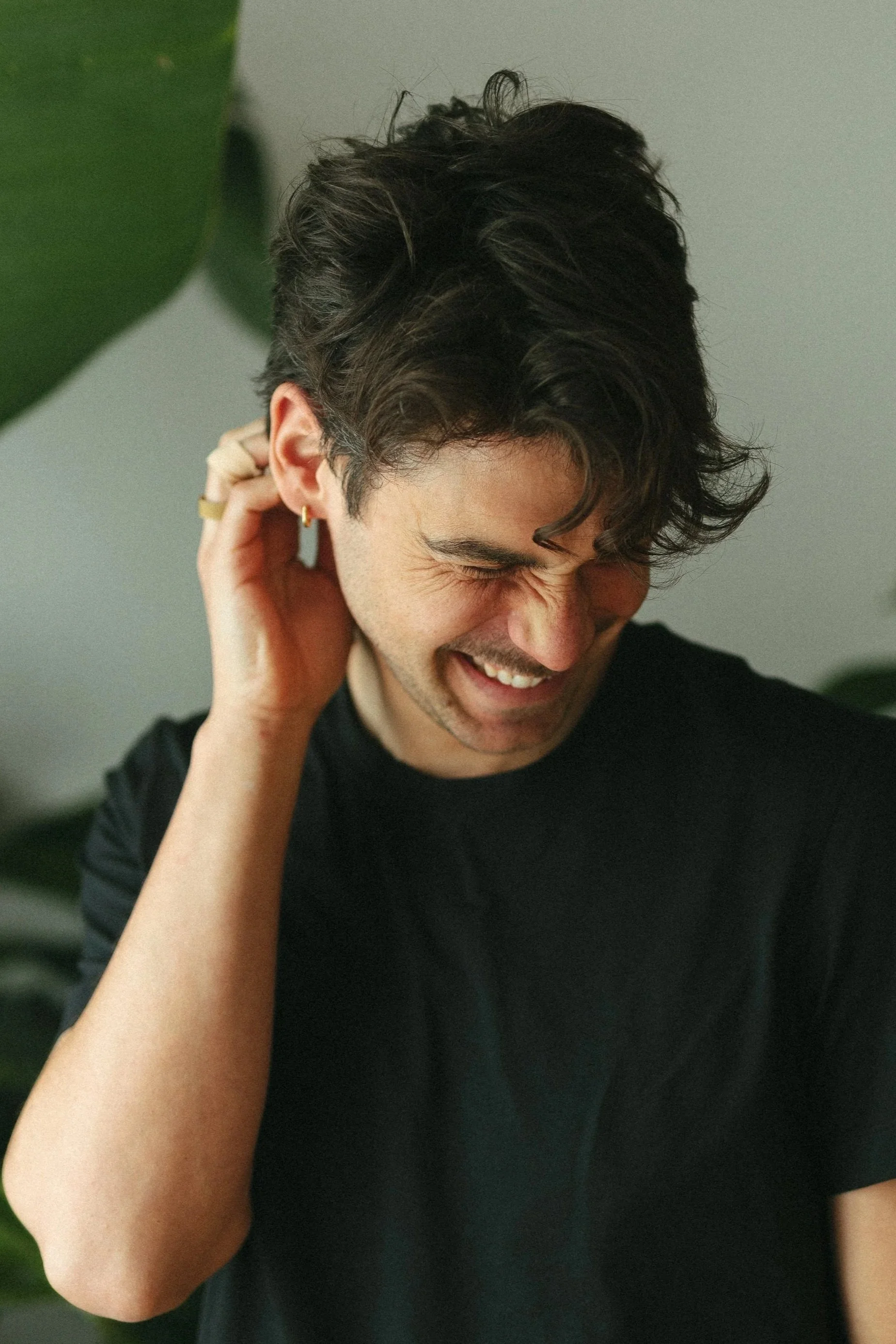 Ein lachender junger Mann mit dunklem, lockigem Haar, der den Kopf leicht neigt, die Augen geschlossen und die rechte Hand hinter dem Ohr hat, vor einem Hintergrund mit grünen Blättern.