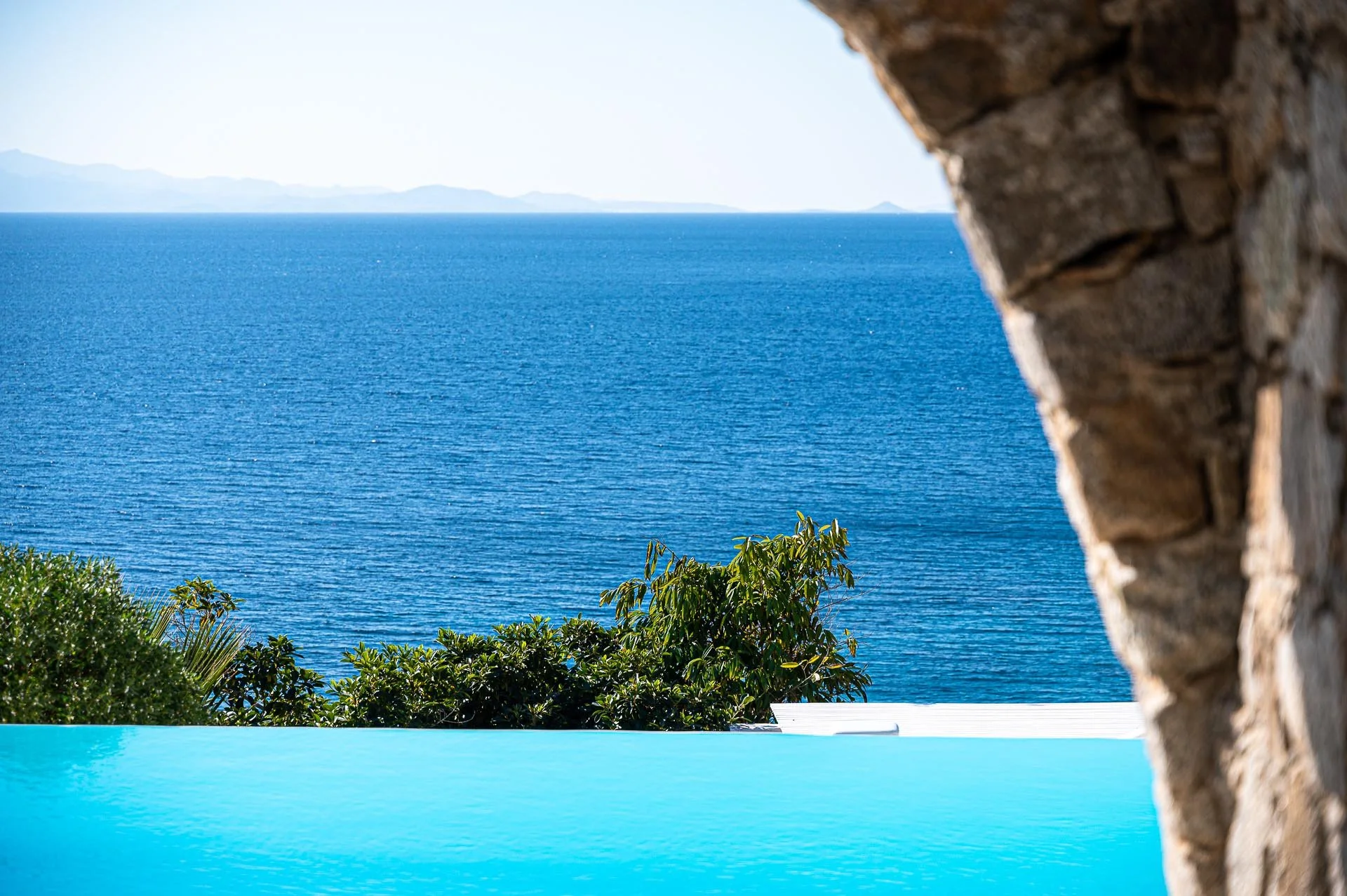 Blick auf den blauen Ozean mit grünen Bäumen im Vordergrund, eine steinige Wand rechts, und eine fernen Insel im Hintergrund.