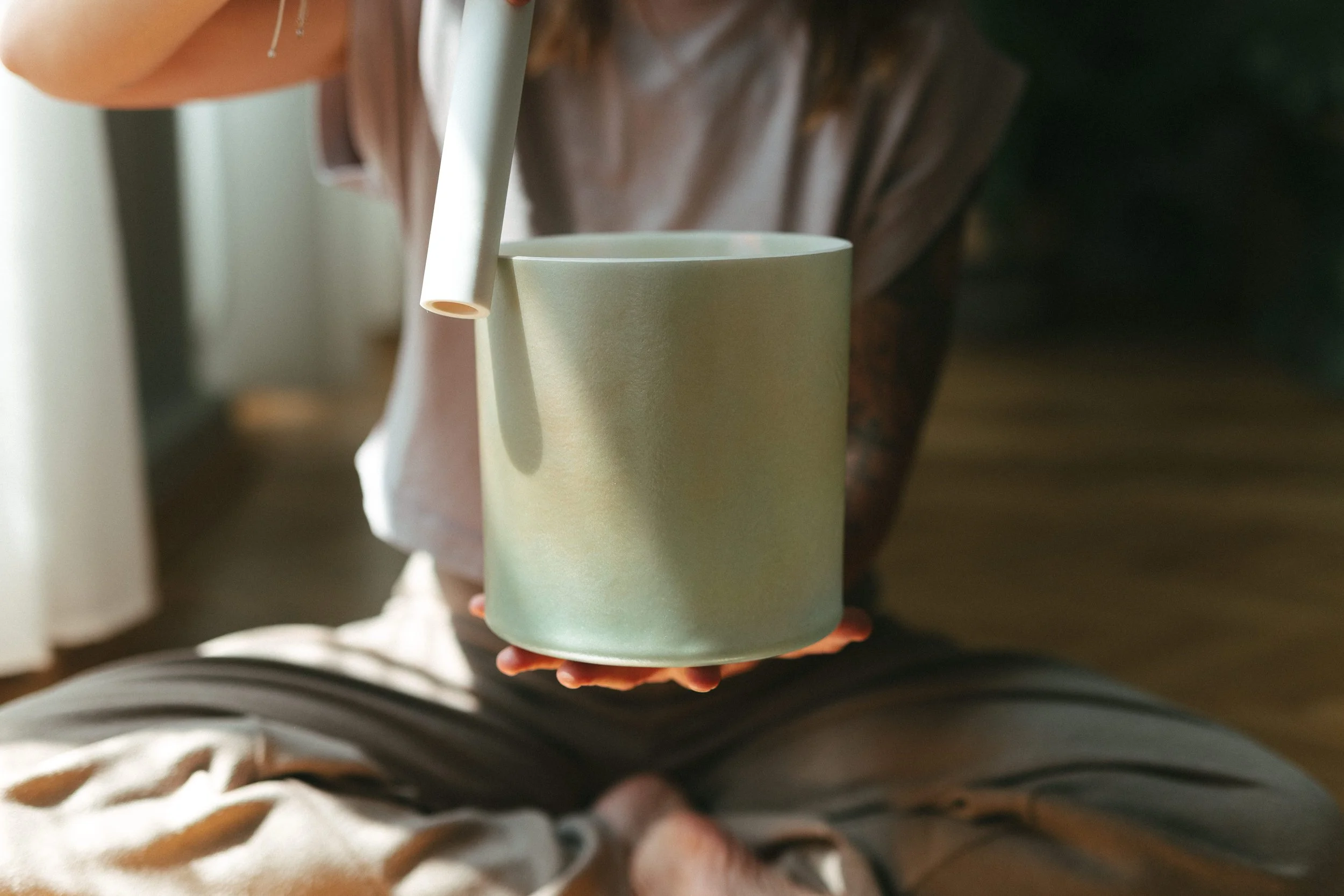 Person hält eine grüne Keramiktasse mit einem weißen Strohhalm in den Händen, sitzend auf einem Boden mit Holzparkett, im Hintergrund Licht durch ein Fenster.