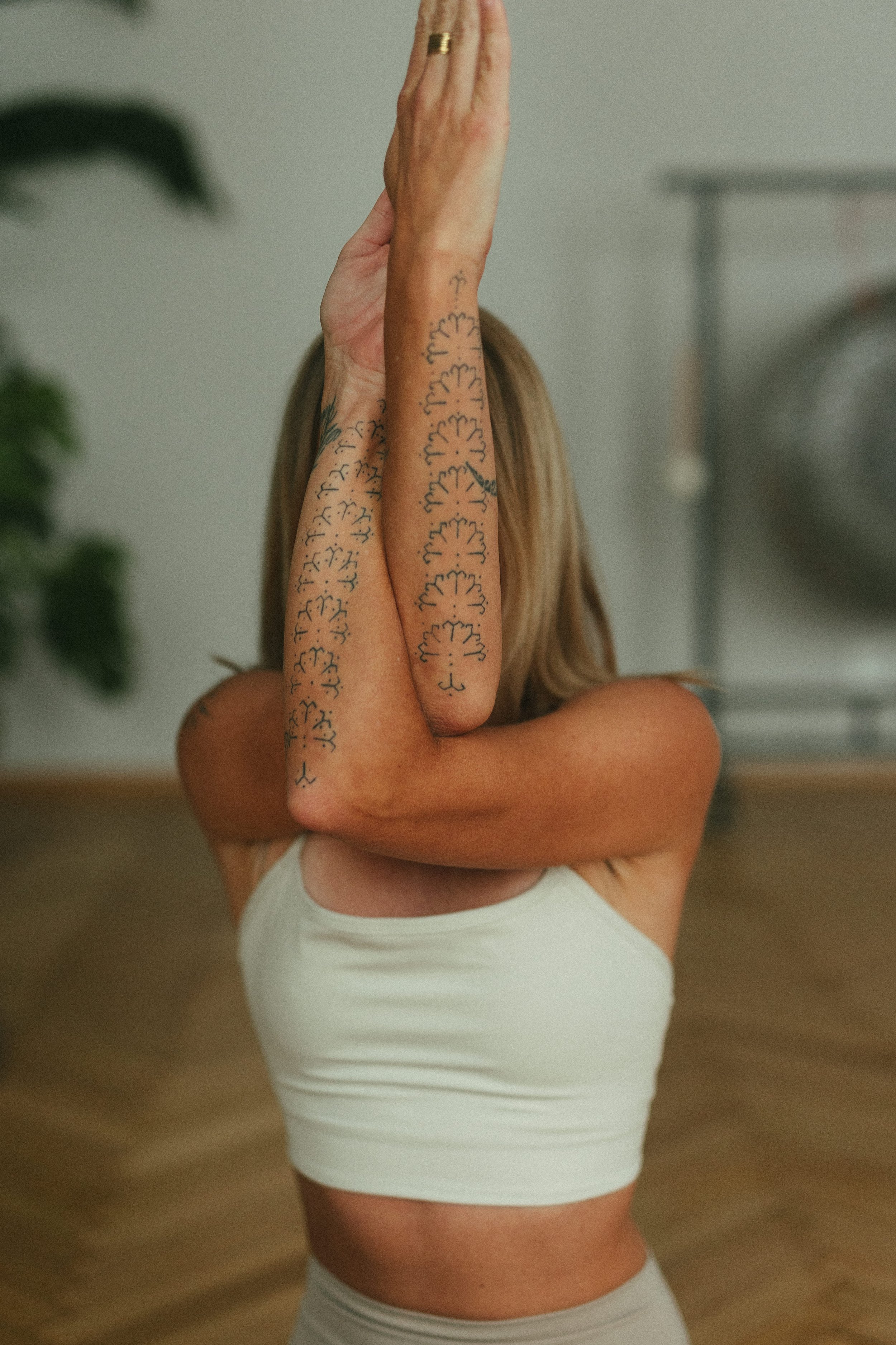 Eine Frau macht eine Yoga-Pose, hebt einen Arm nach oben und legt den anderen Arm quer über den Körper, in einem hellen Raum mit Holzboden und grünen Pflanzen im Hintergrund.