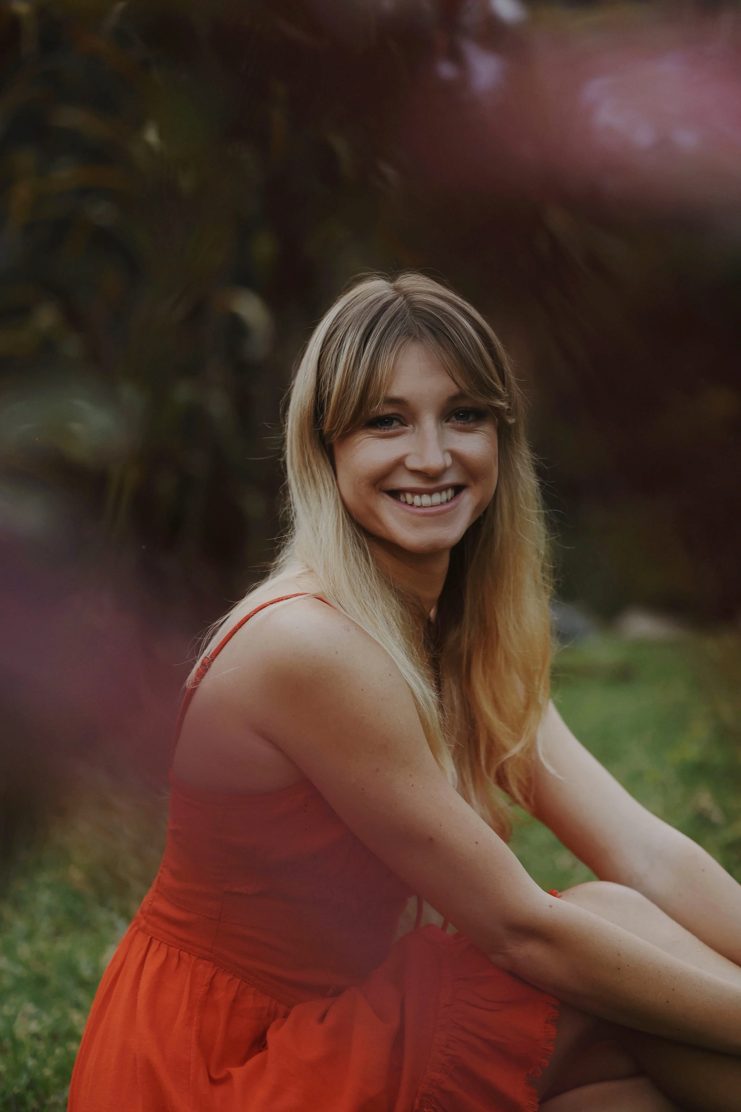 Lächelnde junge Frau mit langen blonden Haaren, trägt ein rotes Kleid und sitzt im Freien auf dem Gras, im Hintergrund Pflanzen und Bäume.