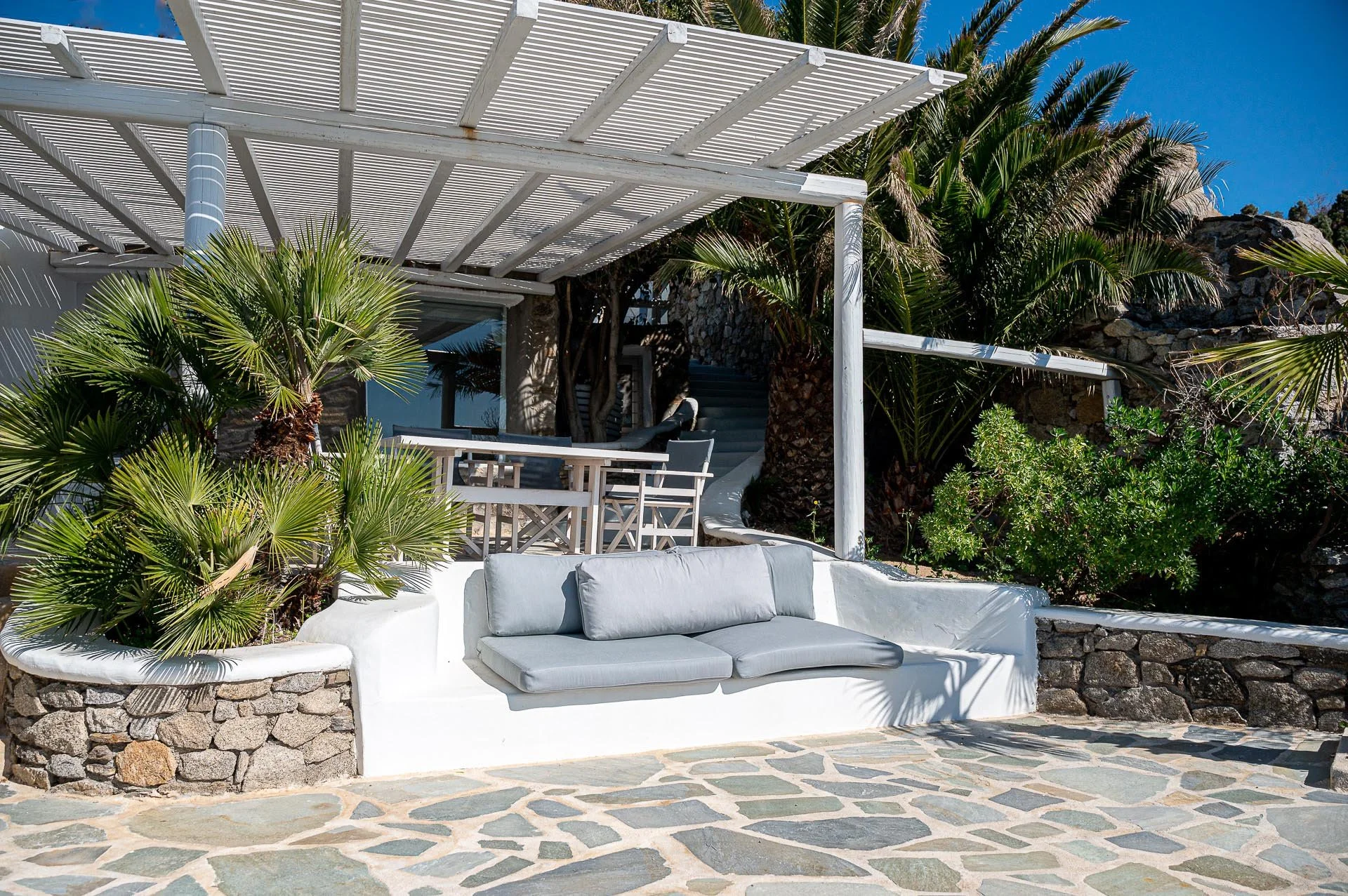 Weiße Gartenlounge mit grauen Kissen, umgeben von Palmen und Steinen, mit einer Pergola im sonnigen Außenbereich