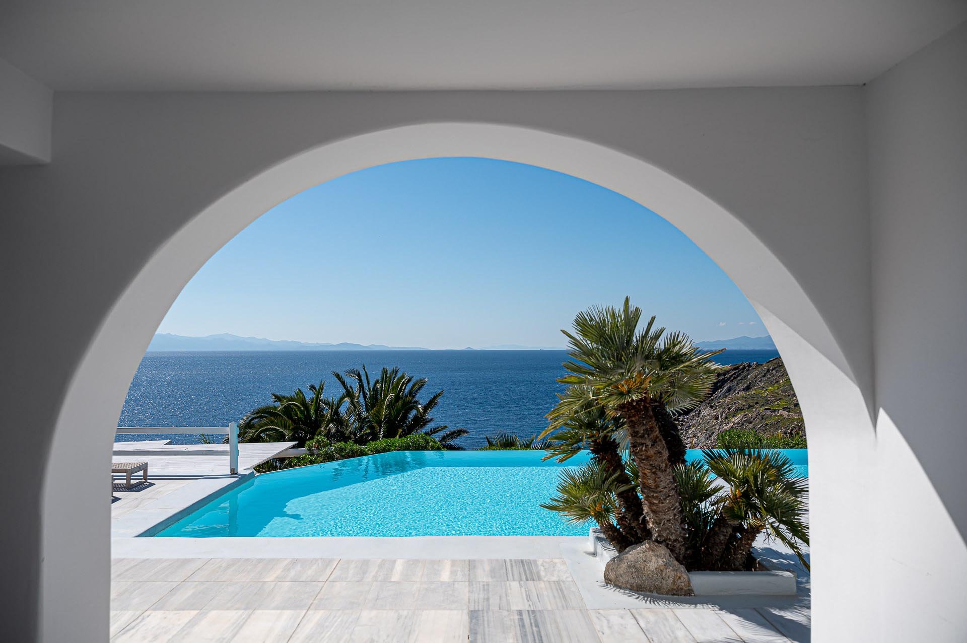 Blick durch eine weiße Archöffnung auf einen Infinity-Pool mit Blick auf das Meer und den Himmel, umgeben von Palmen auf einer Terrasse