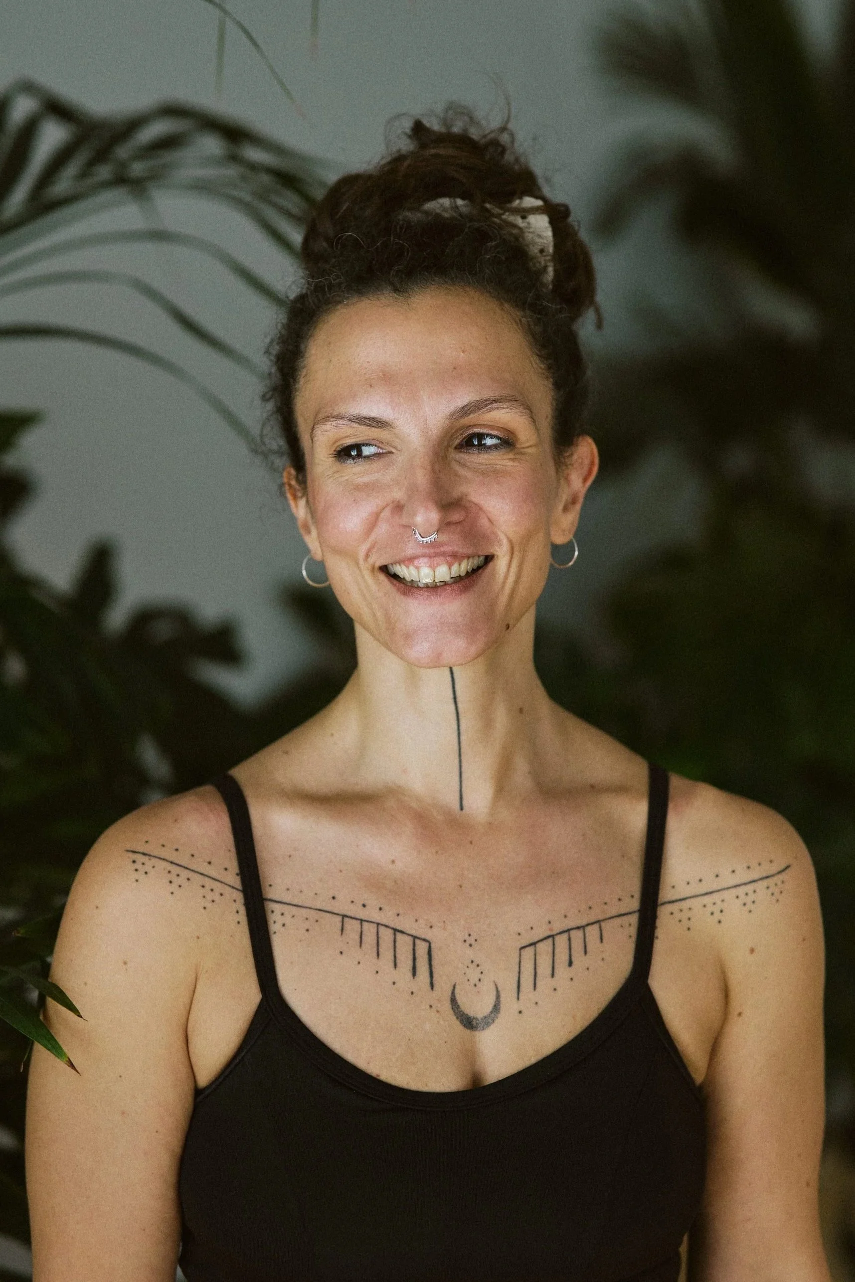Eine Frau mit dunklem, lockigem Haar und tatooierter Schulter in Seitenansicht, die nach unten blickt, im Hintergrund mit grünen Blättern und hellem Licht.