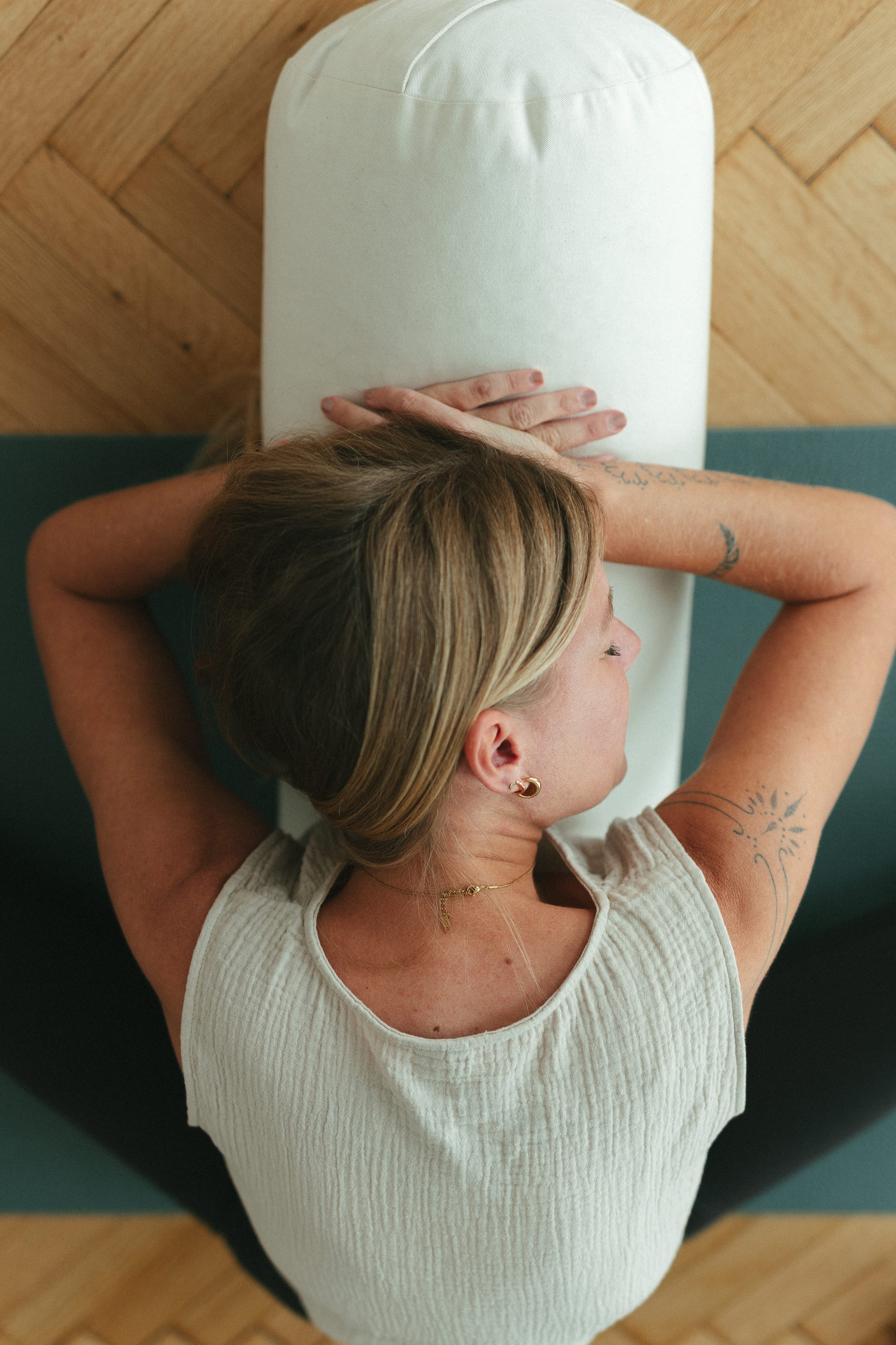 Frau liegt auf einer Yoga-Matte, entspannt mit geschlossenen Augen, Arme hinter dem Kopf, auf dem Rücken liegend, im Raum mit Holzboden.
