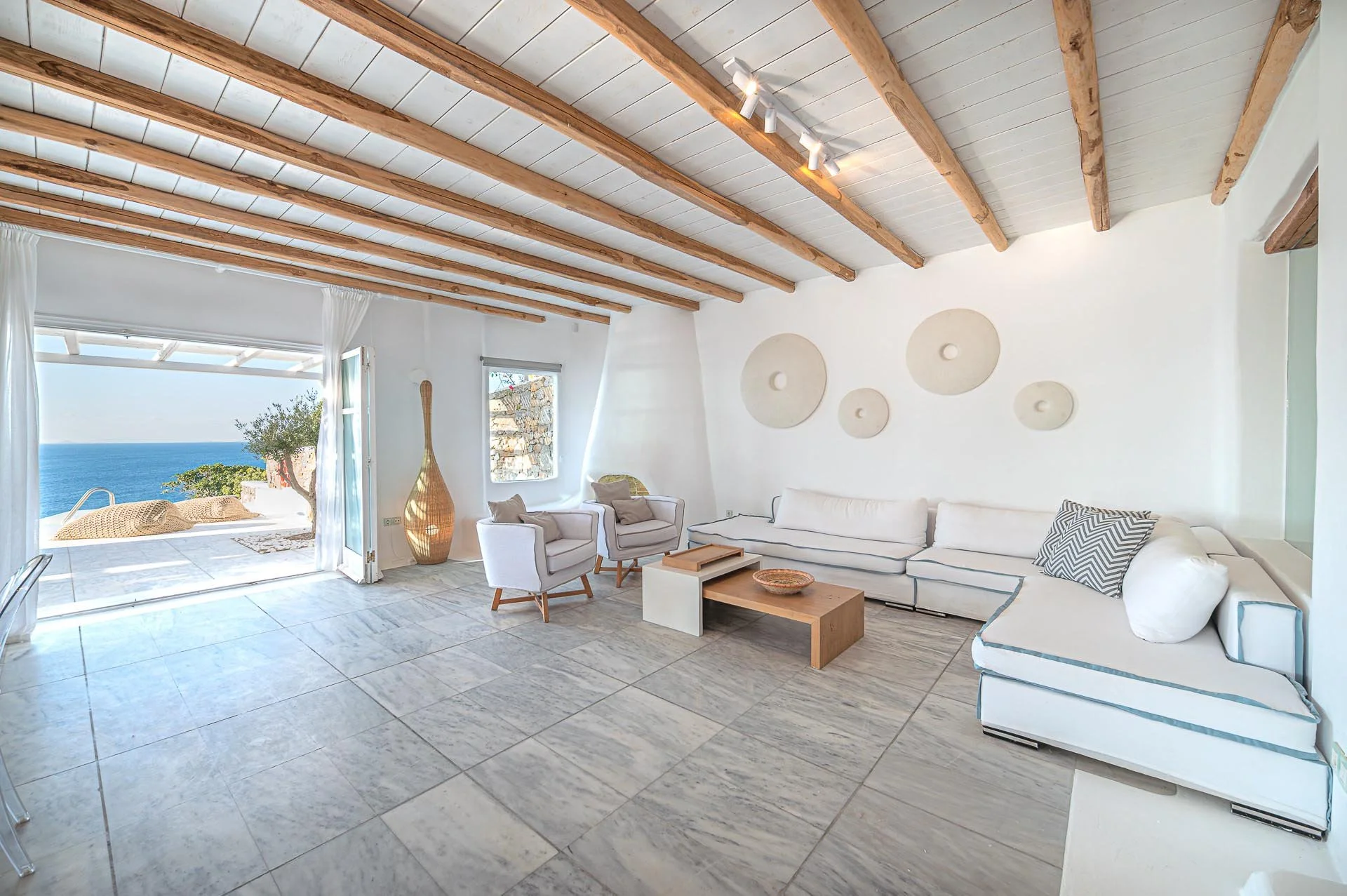Wohnzimmer mit weißen Möbeln, Blick auf das Meer durch offene Terrassentür, Holzbalkendecke, Wanddekorationen