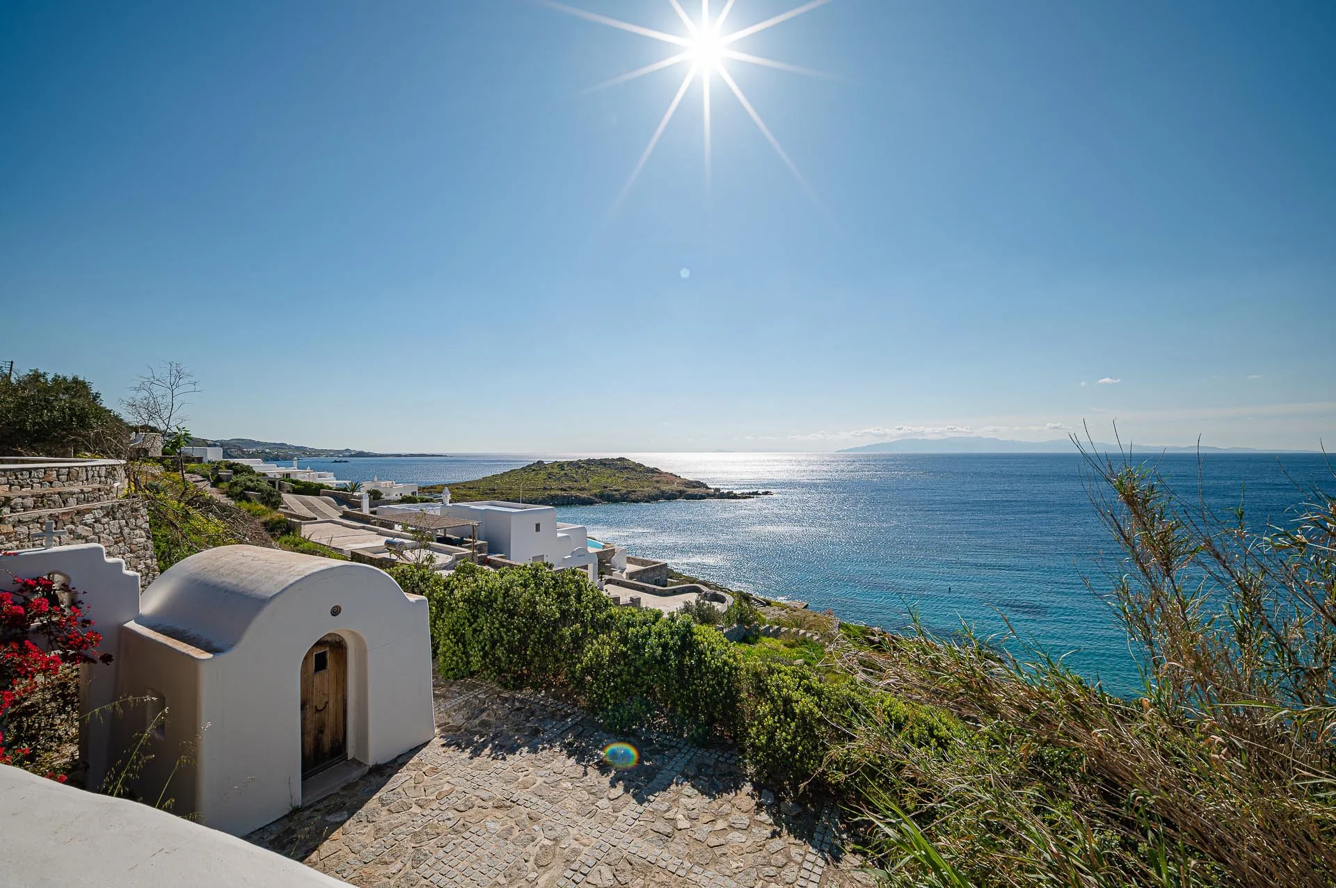 Weiße Häuser auf einem Hügel mit Blick auf das Meer unter klarem blauen Himmel, herum mit grünen Pflanzen und Steinen.