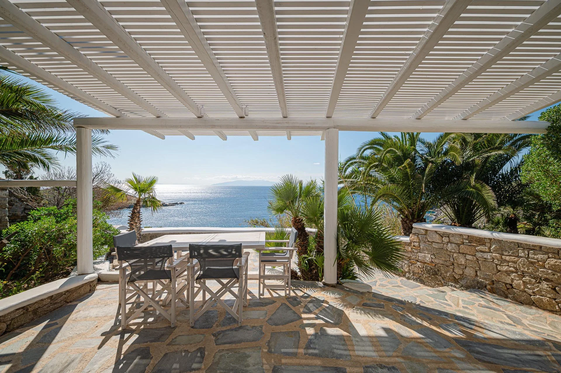 Blick auf eine Terrasse mit einem Esstisch und Stühlen, umgeben von Palmen, mit Blick auf das Meer und den Himmel im Hintergrund.