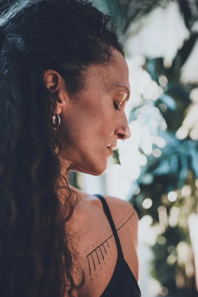 Eine Frau mit dunklem, lockigem Haar und tatooierter Schulter in Seitenansicht, die nach unten blickt, im Hintergrund mit grünen Blättern und hellem Licht.