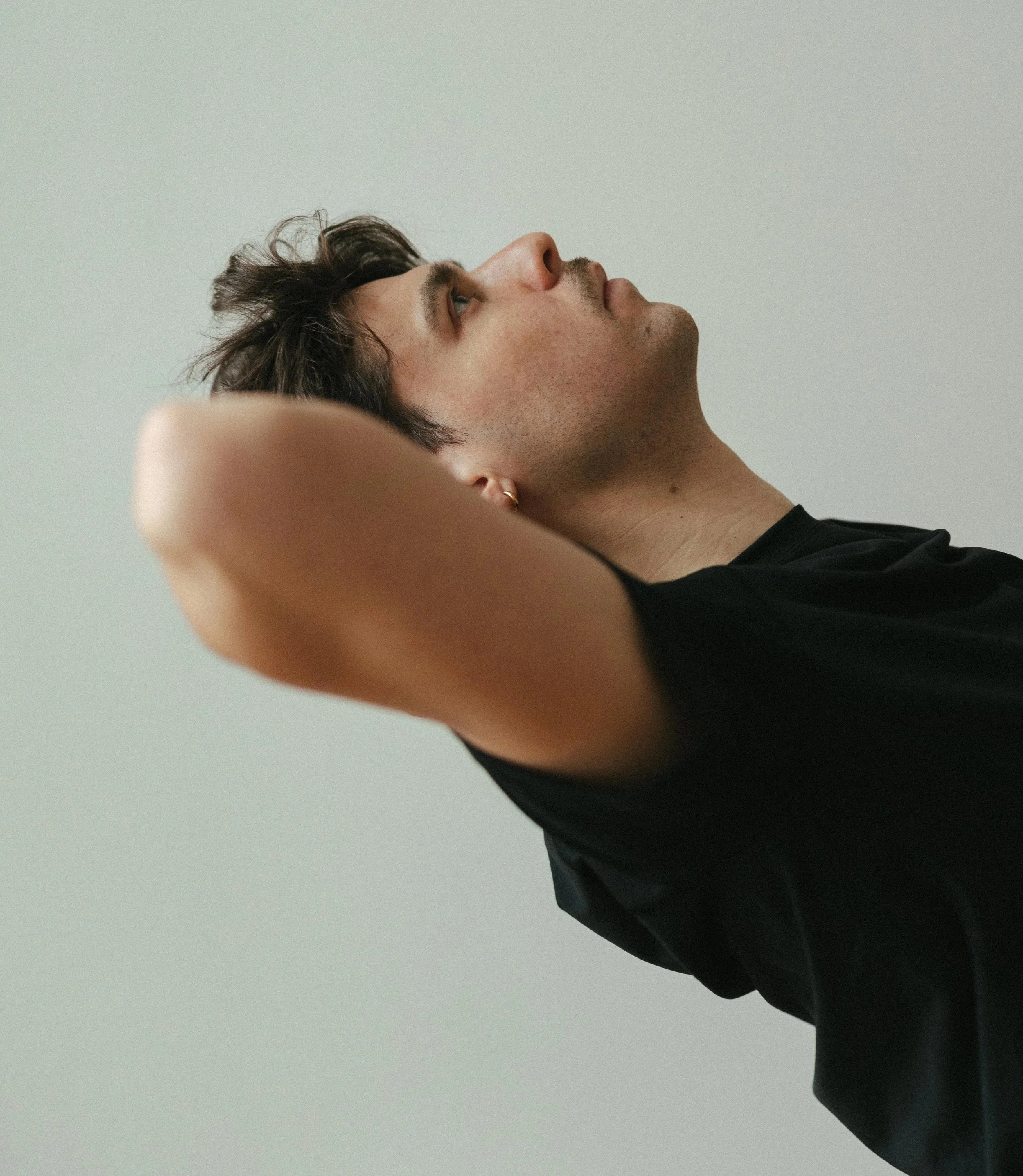 Junge Person mit dunklem Haar, trägt ein schwarzes T-Shirt, hält die Arme hinter dem Kopf, blickt nach oben, neutraler Hintergrund.