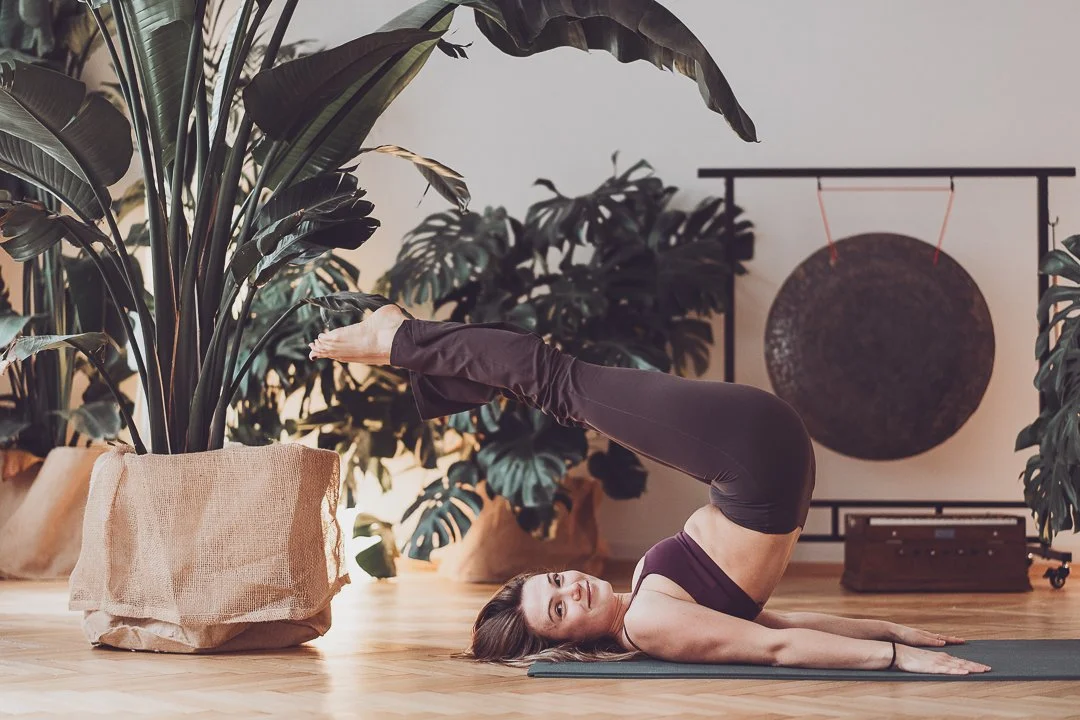 Frau macht Yoga in einem Raum mit vielen Pflanzen, liegend auf dem Rücken mit den Beinen nach oben gegen eine Pflanze gestützt.