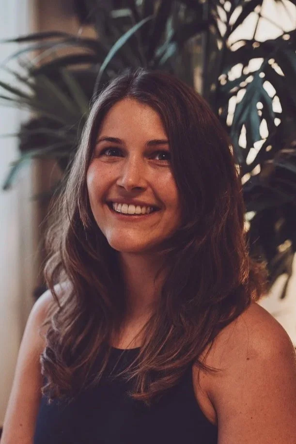 Ein Lächelndes junges Frau mit langen, braunen Haaren vor einer Pflanzenkulisse.