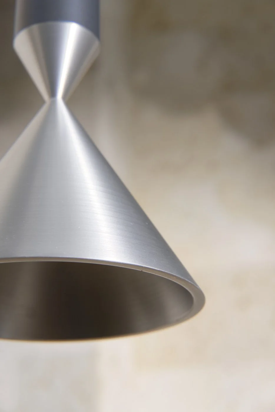 Close-up of a metallic cone-shaped object, likely a lampshade, with a smooth brushed finish and a hollow interior, against a blurred neutral background.