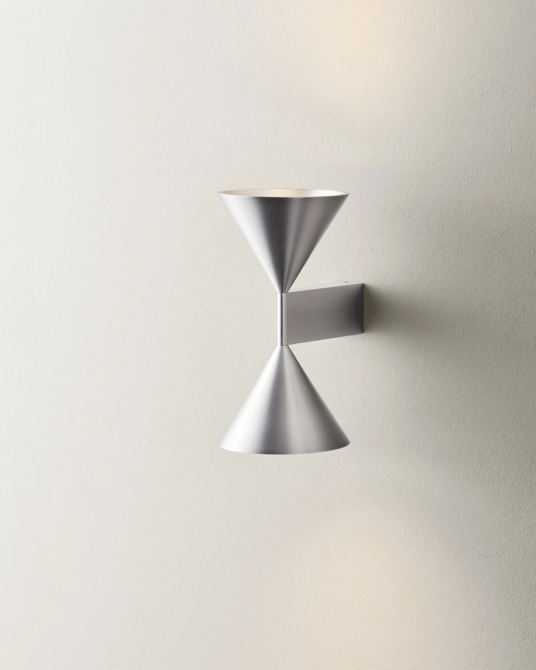 Modern wall sconce with two conical metal shades mounted on a beige wall, casting reflections.