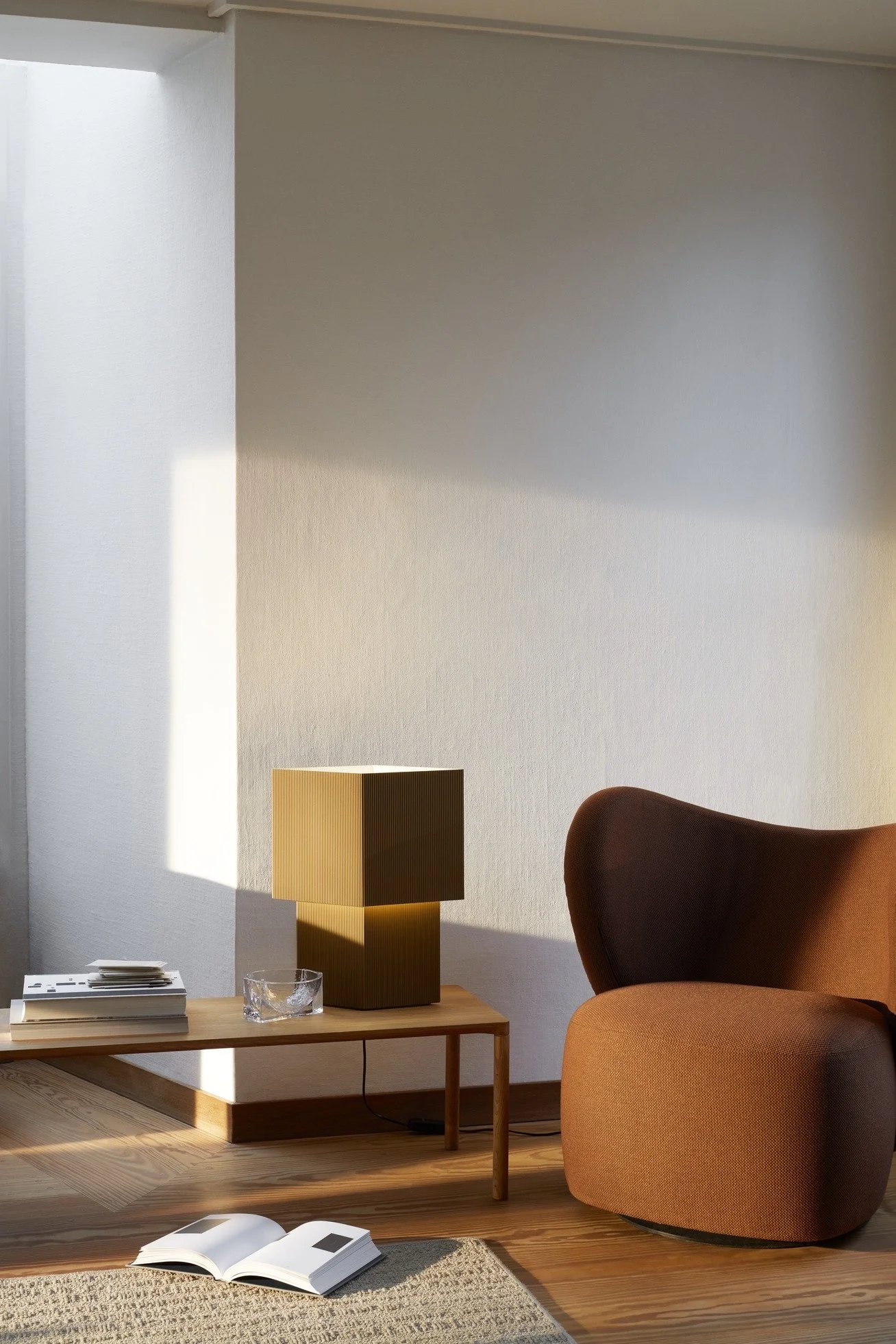 Minimalist living room corner with a brown armchair, wooden side table with a stack of books, a glass ashtray, and a modern table lamp. Sunlight casts a shadow on the wall and floor.