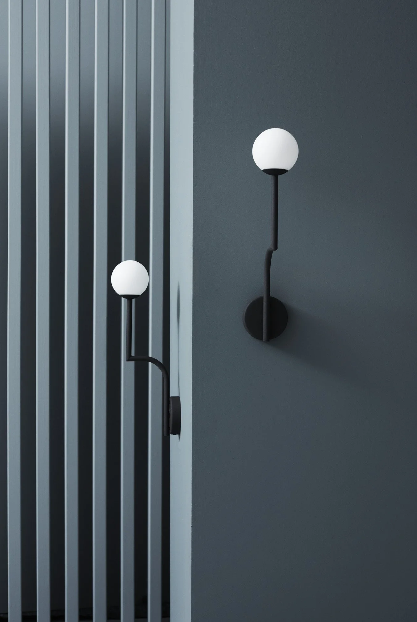 Two modern wall-mounted black lamps with white globe-shaped bulbs on a dark gray wall, next to a metallic slatted surface.