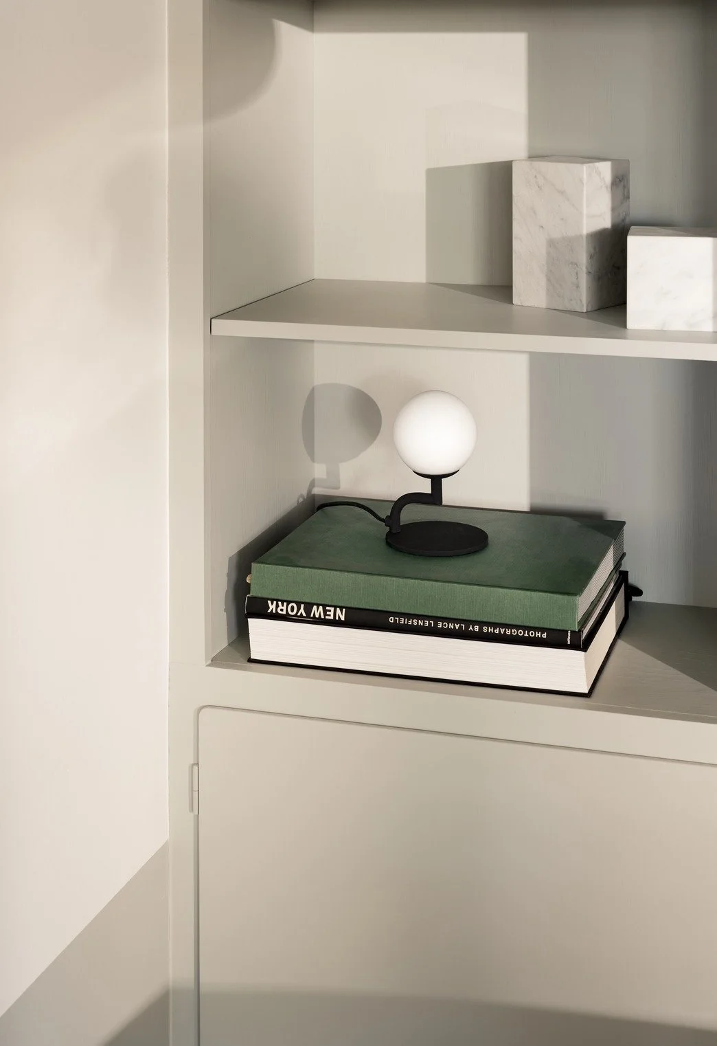 A white shelf with three marble blocks on the top and a small black and white lamp on two large books, one green and one black, on the bottom shelf.
