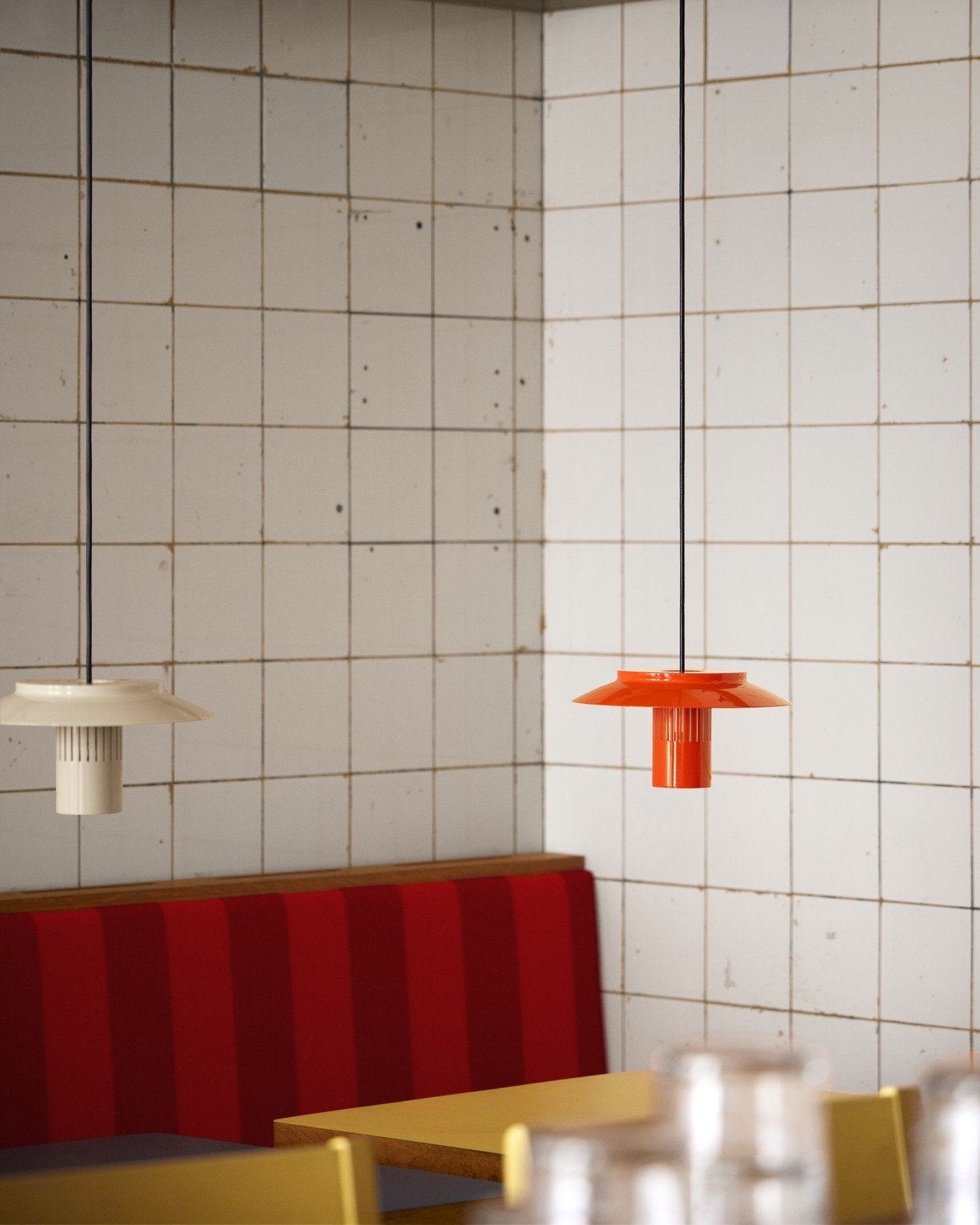 Interior of a restaurant or cafe with white tiled walls, a yellow table, red and yellow chairs, and two pendant lights, one white and one orange, hanging from the ceiling.