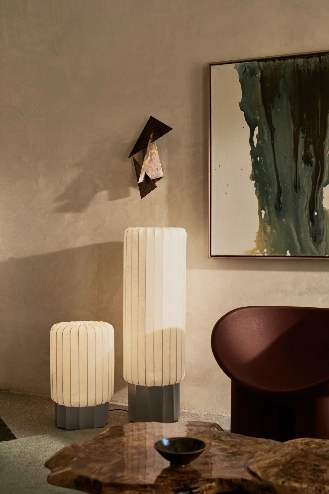 A modern interior wall with a metallic abstract sculpture, a large abstract painting, two tall upholstered ottomans, a dark curved chair, and a wooden table with a bowl