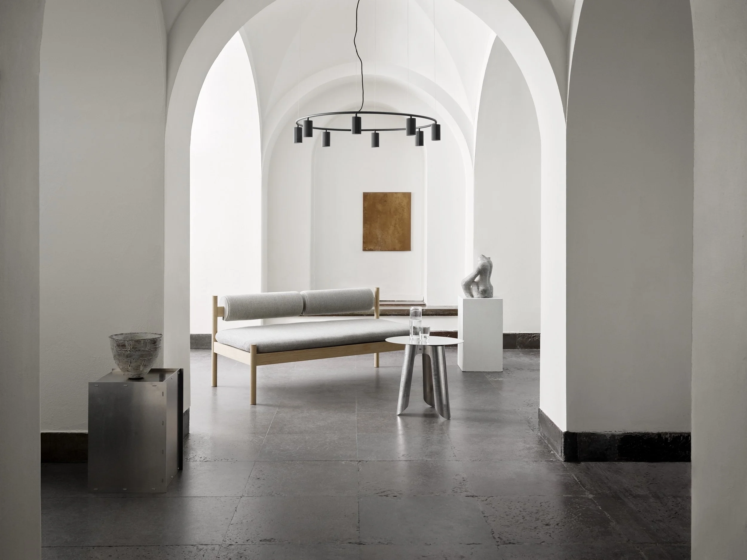 Minimalist modern interior with white arched walls, a light gray sofa with wooden legs, a small white round table with water and glasses, a bronze-colored wall art, a sculpture on a white pedestal, and other decorative objects.