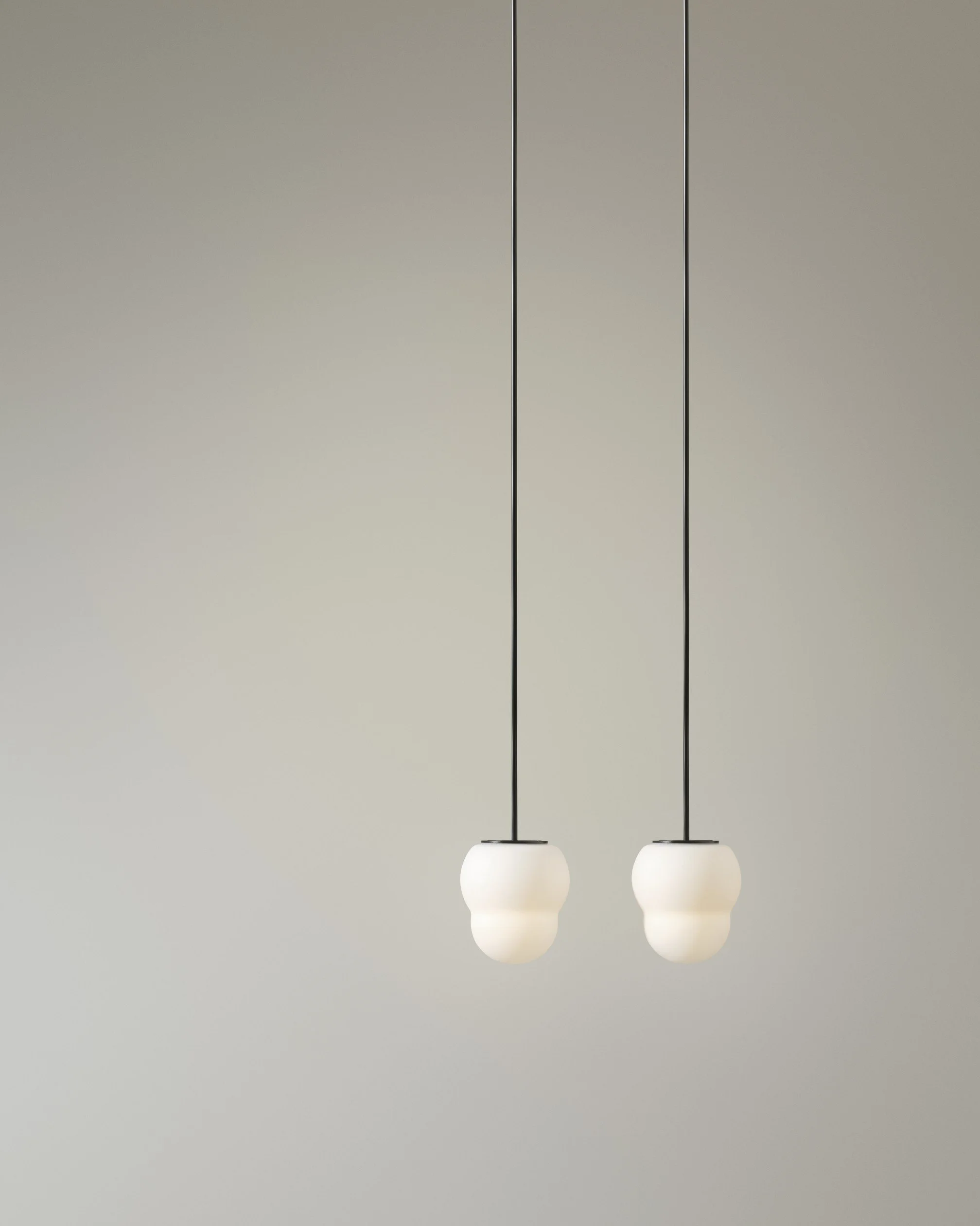 Two pendant lights with white rounded shades hanging from a ceiling with black cords over a plain wall.