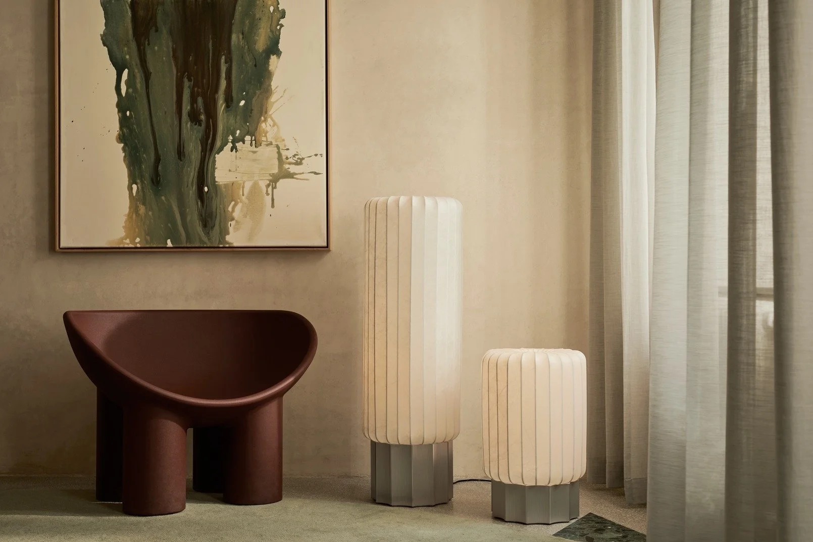 Interior of a living room with abstract wall art, a rounded brown chair, and two tall, white, ribbed lamps on rectangular bases near a window with sheer curtains.