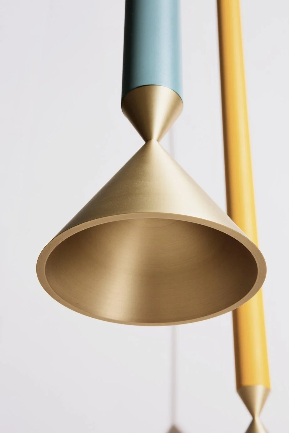 Close-up of a modern hanging lamp with a gold conical shade, a teal stem, and a yellow pencil-shaped element in the background.