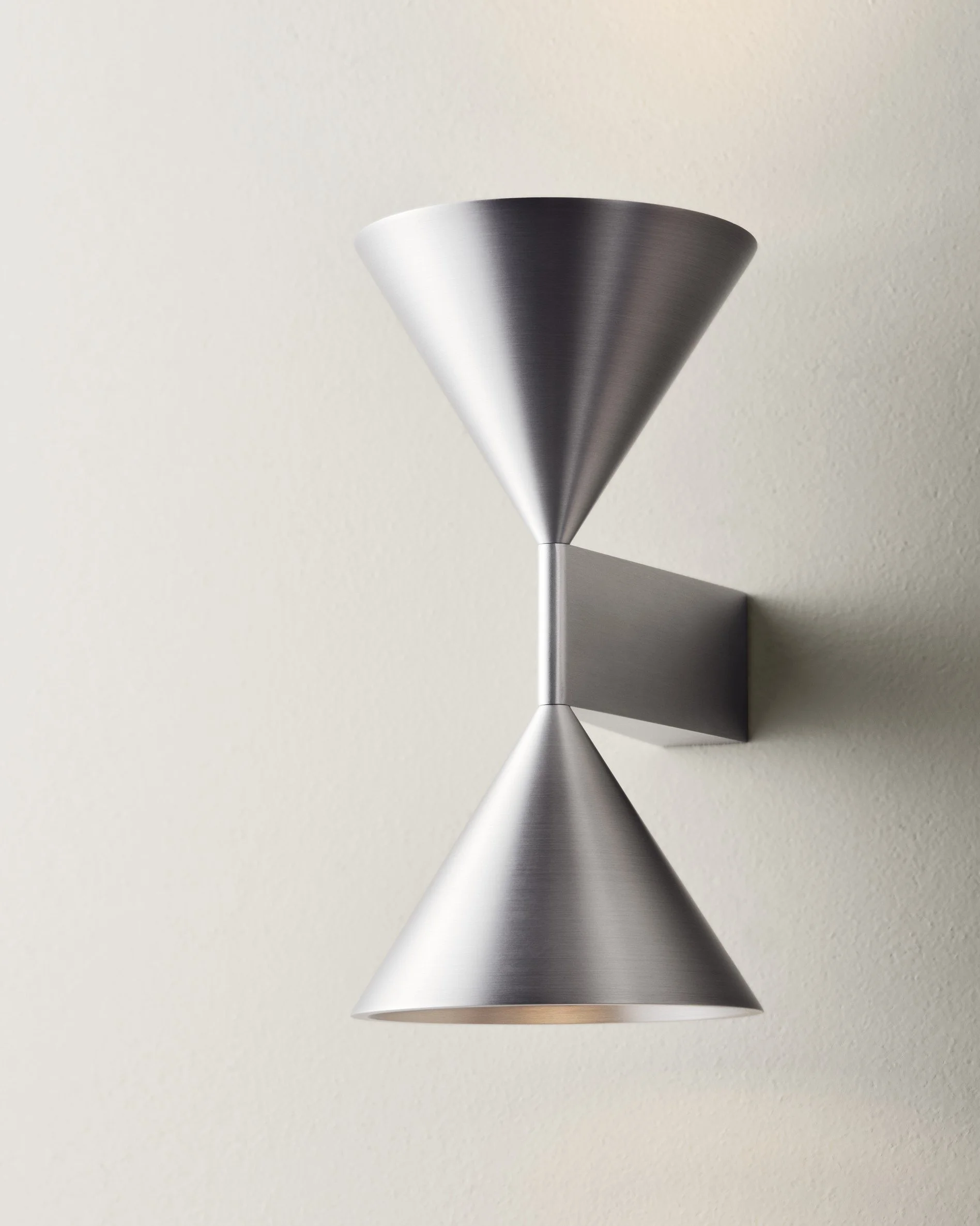 Modern wall-mounted light fixture with two conical shades, one pointing up and one pointing down, made of brushed metal, mounted on a plain light-colored wall.