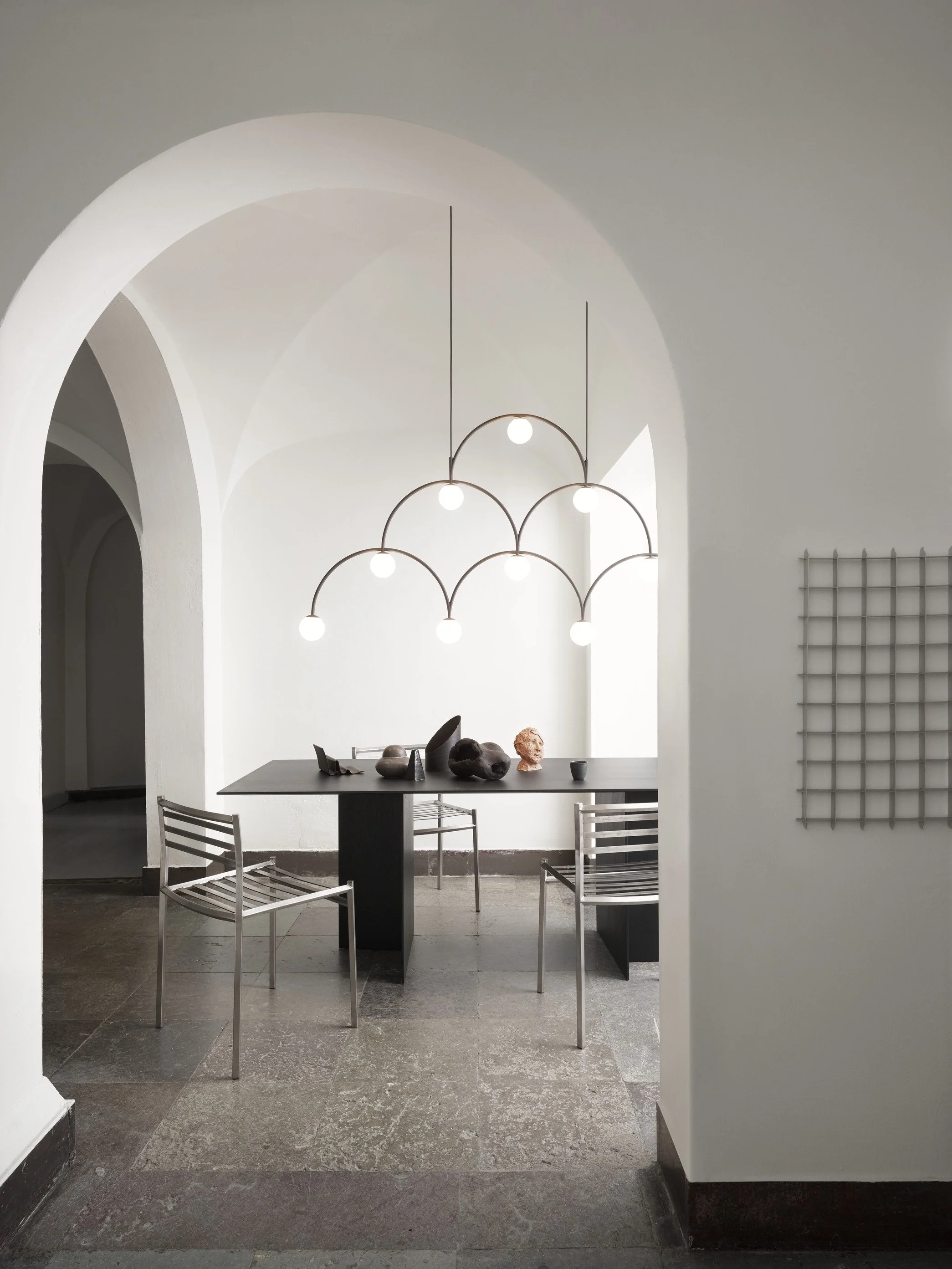Modern dining area with a black table, metal chairs, and an artistic chandelier with multiple globe lights, minimalist decor, white walls, and a tiled floor.
