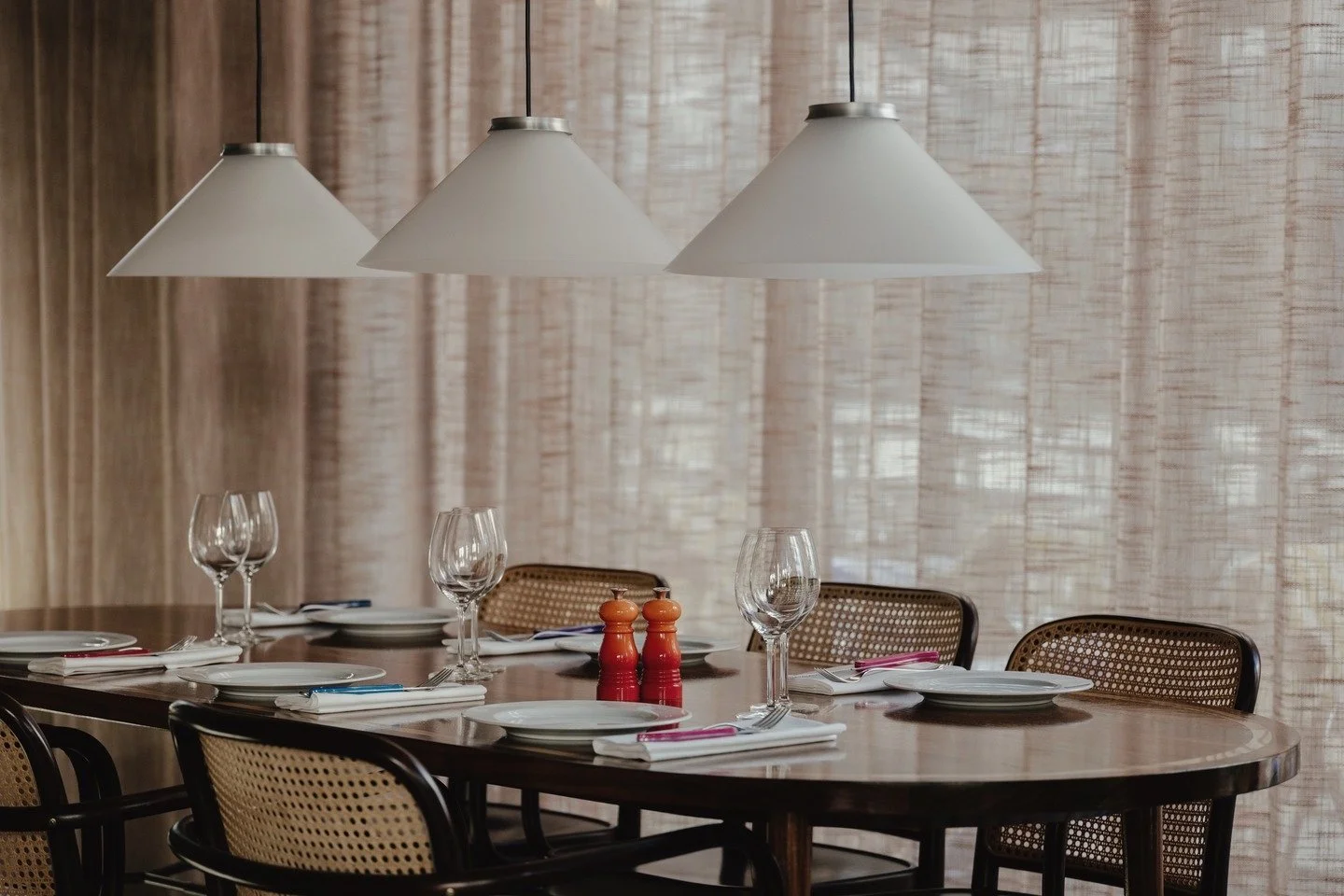Empty dining table set with plates, wine glasses, napkins, and salt and pepper shakers in a room with sheer curtains and hanging pendant lights.