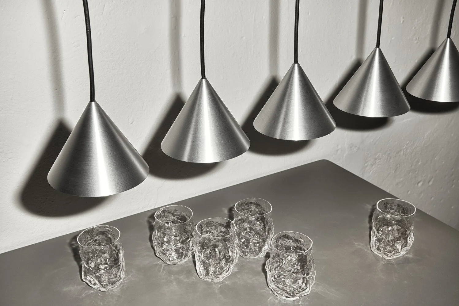 Five metallic cone-shaped pendant lights hanging above a gray surface with six textured clear drinking glasses filled with water.