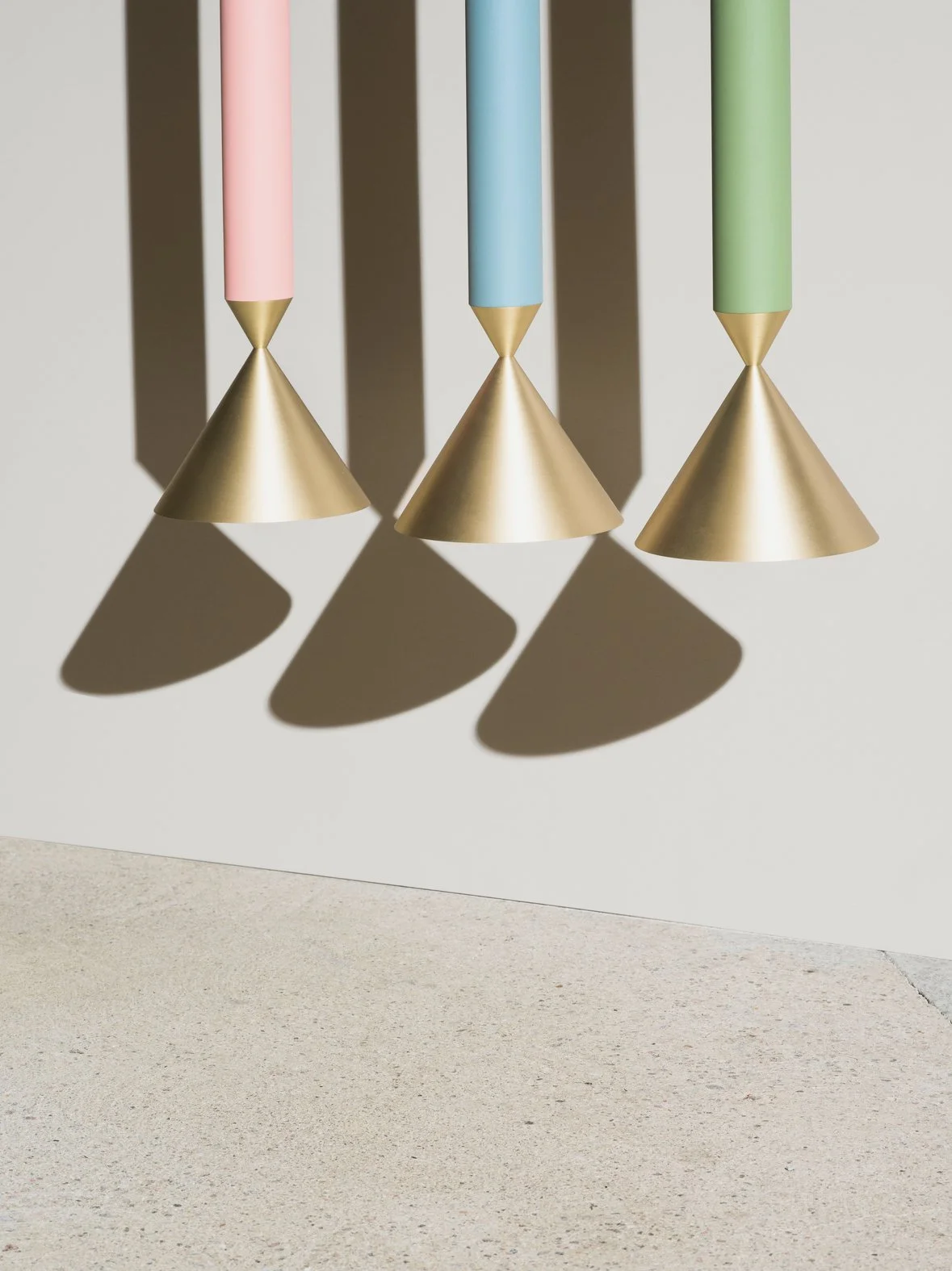 Three hanging objects with pink, blue, and green handles, cone-shaped gold bases, and casting shadows on a light-colored surface.