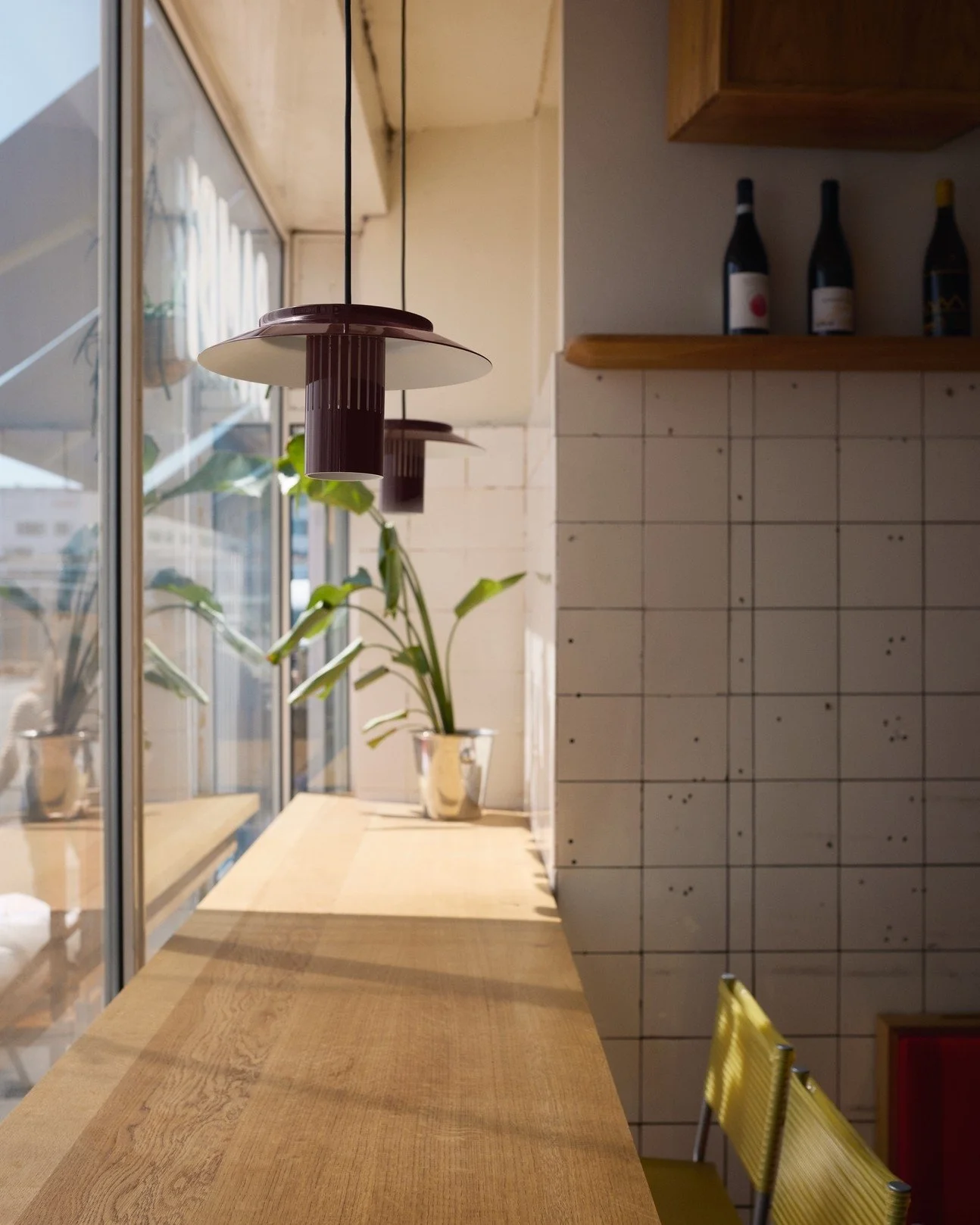 Interior view of a cozy café with a wooden counter, hanging pendant lights, potted plant, and a tiled wall with bottles on a shelf, illuminated by natural light from large windows.