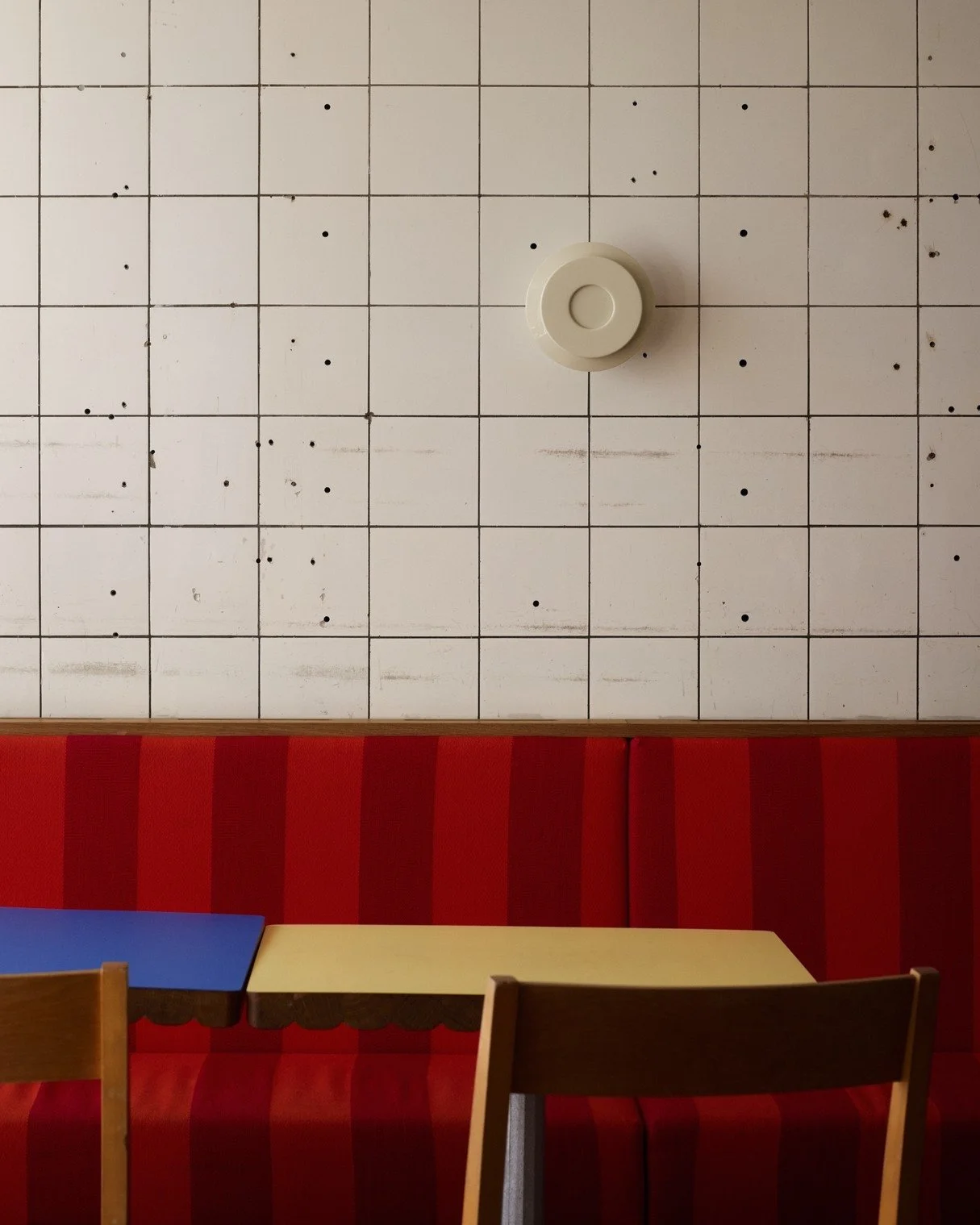 A white tiled wall with black dots, a white circular thermostat, and a red cushioned bench with wooden tables and chairs in front.