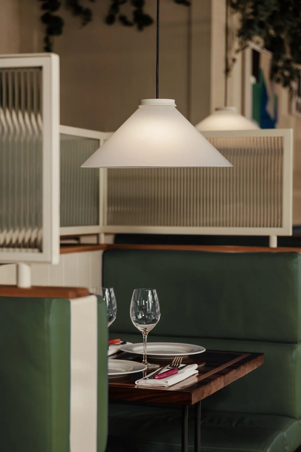 Empty restaurant booth with a wooden table set for two, including wine glasses, plates, silverware, and napkins, under hanging white pendant lights.