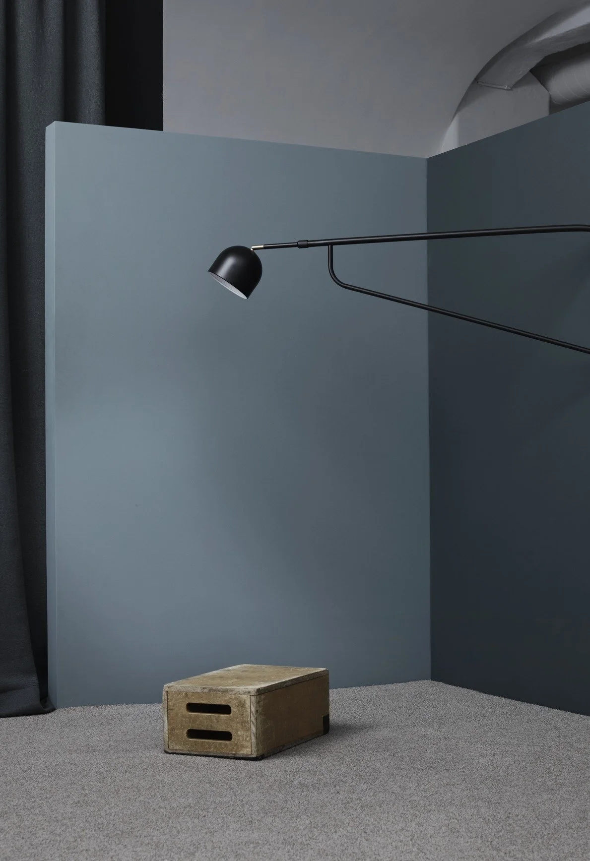 Minimalist interior with a wooden box on a gray carpet, dark curtain, and a modern black wall-mounted lamp extending over a blue wall.