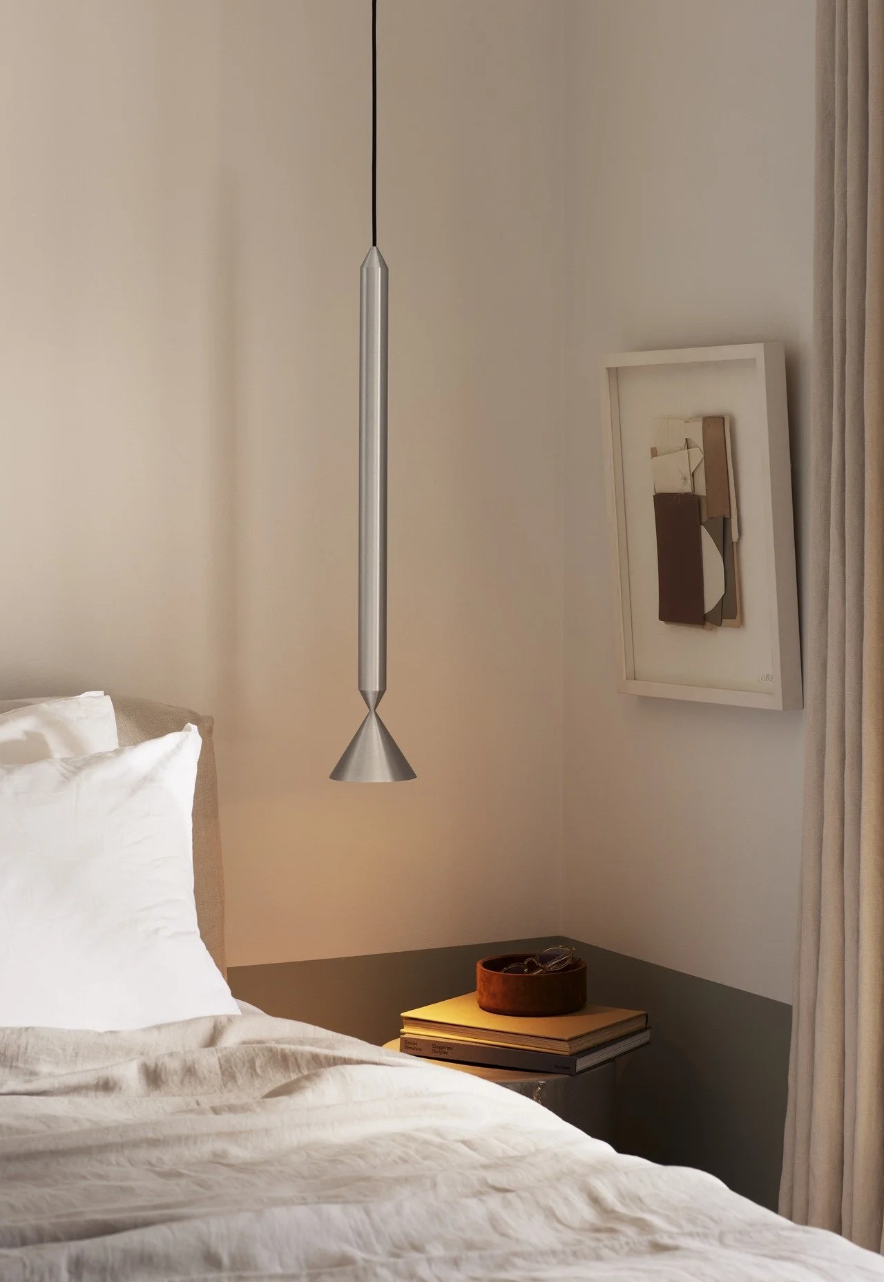 Modern bedroom with a beige headboard, white bedding, a nightstand with books and a decorative bowl, a framed abstract art piece, and a ceiling pendant light.