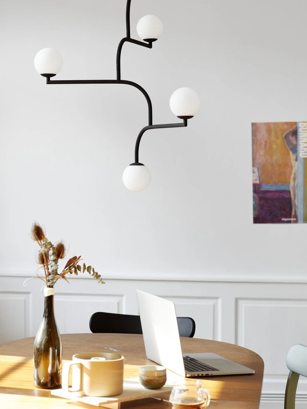 A dining room with a wooden table, a vase with dried flowers, a laptop, and mugs, with a modern black and white chandelier and a colorful abstract painting on the wall.