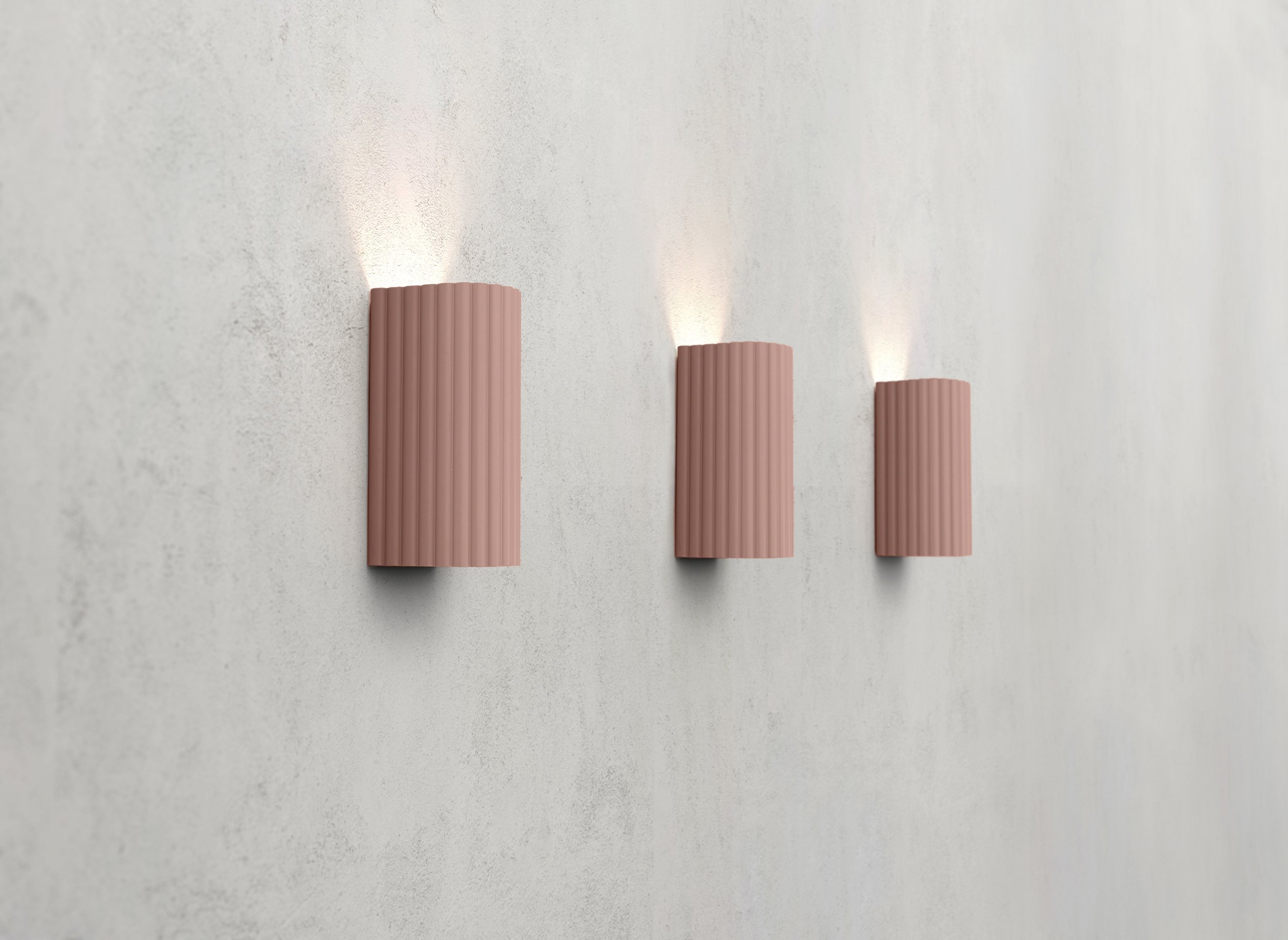 Three pink, vertically ridged wall sconces emitting light onto a white textured wall.