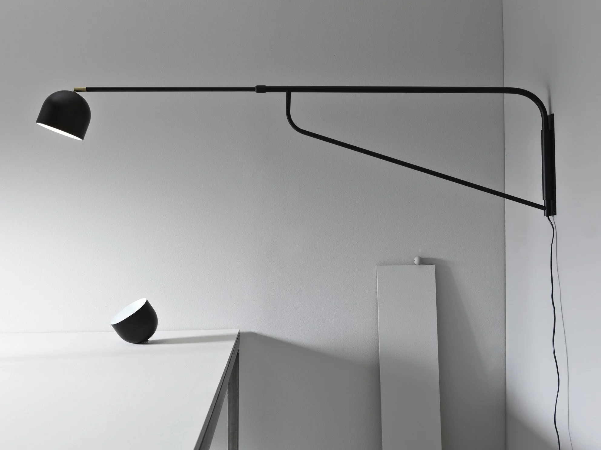 Black wall-mounted lamp directed toward a white table with a black and white mug on it, in a minimalist room with white walls and gray accents.