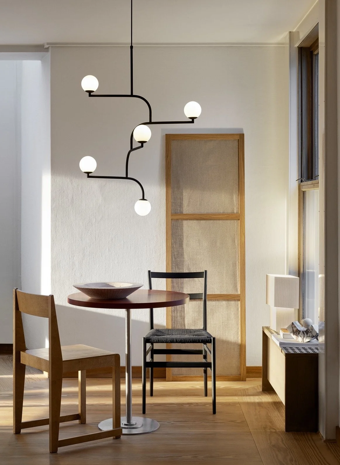 Interior of a room with a round wooden table, three chairs, a modern black and white hanging light fixture, a large window, a wooden framed room divider, and a small bench with a white lamp and decorative object.