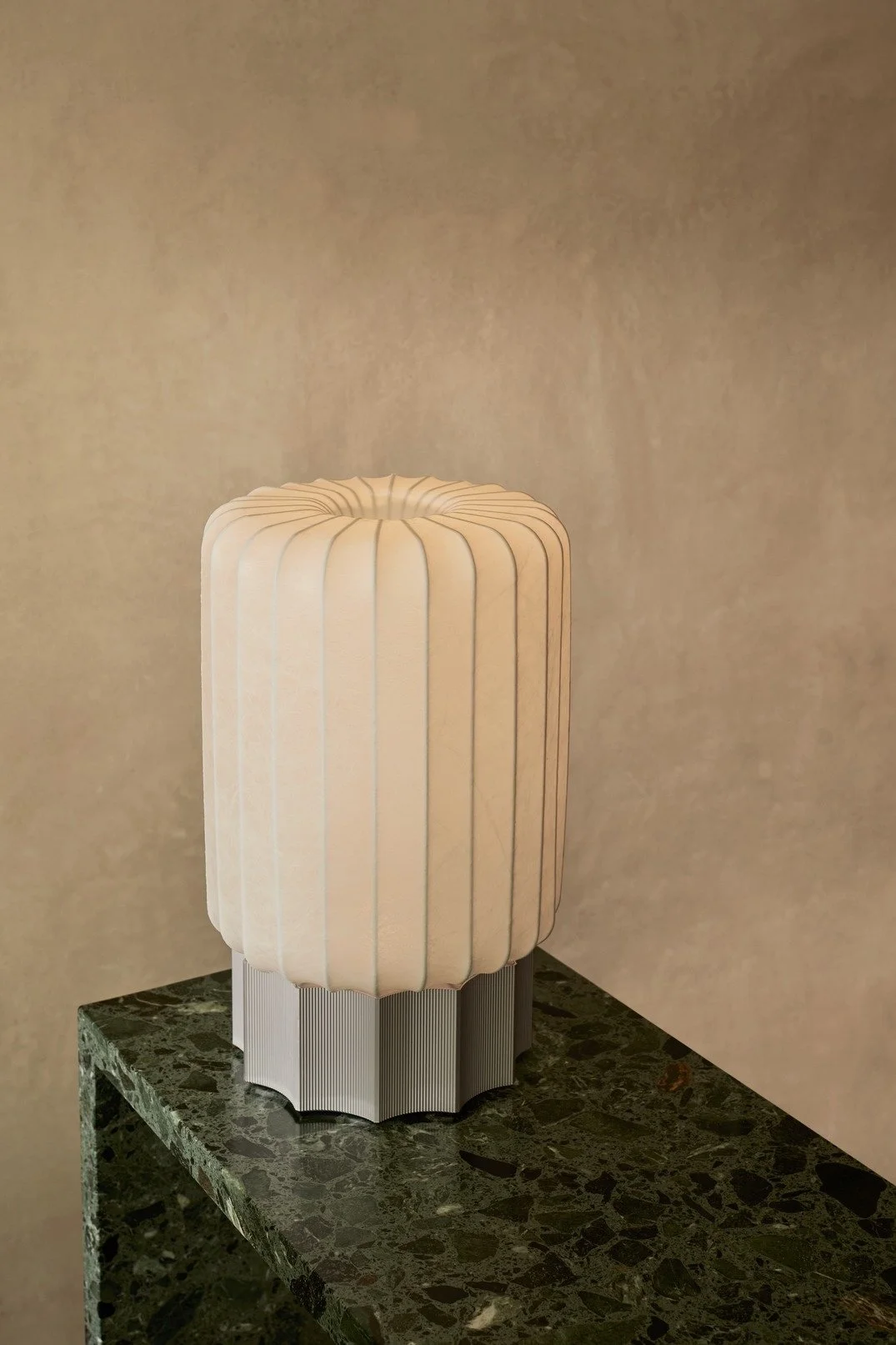 A table lamp with a pleated white shade and a round base, placed on a dark green marble table against a beige wall.