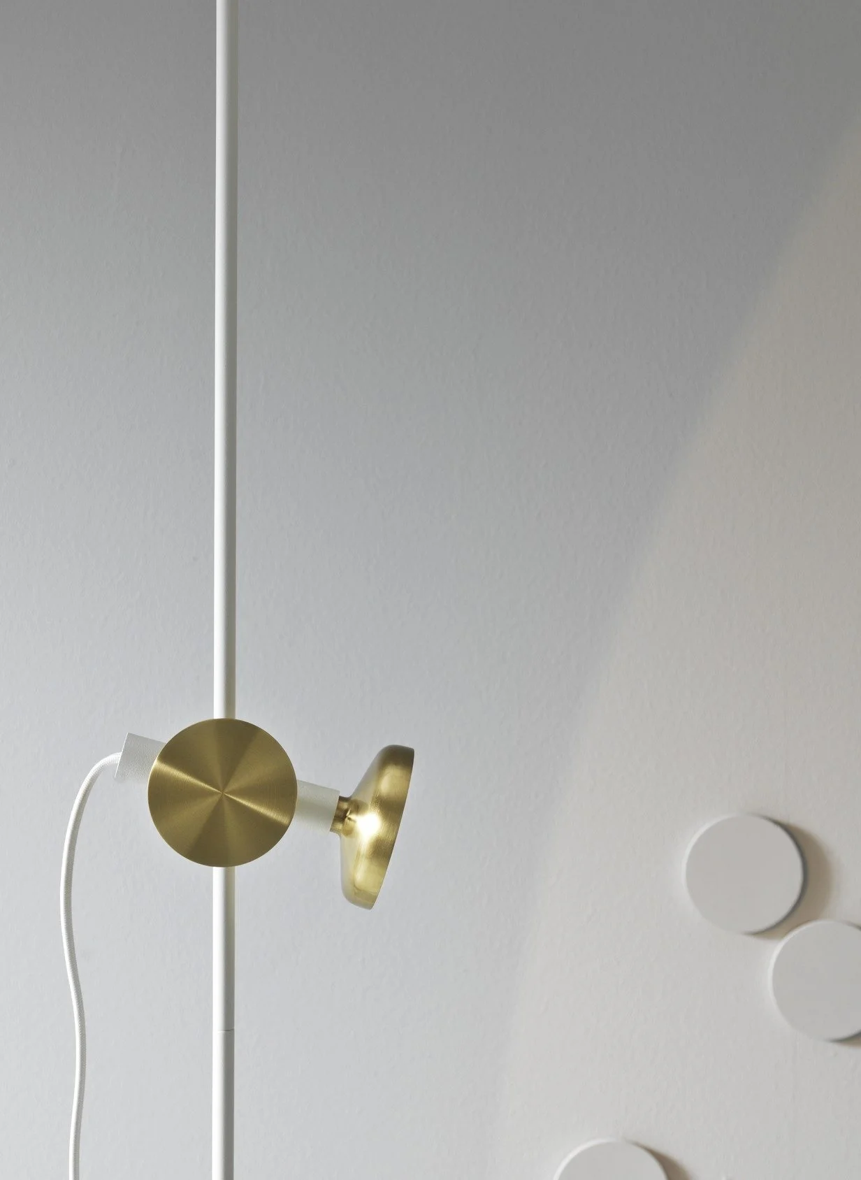 Close-up of a modern white and gold wall-mounted lamp with a round gold shade, attached to a white pole, with a plain white wall behind.
