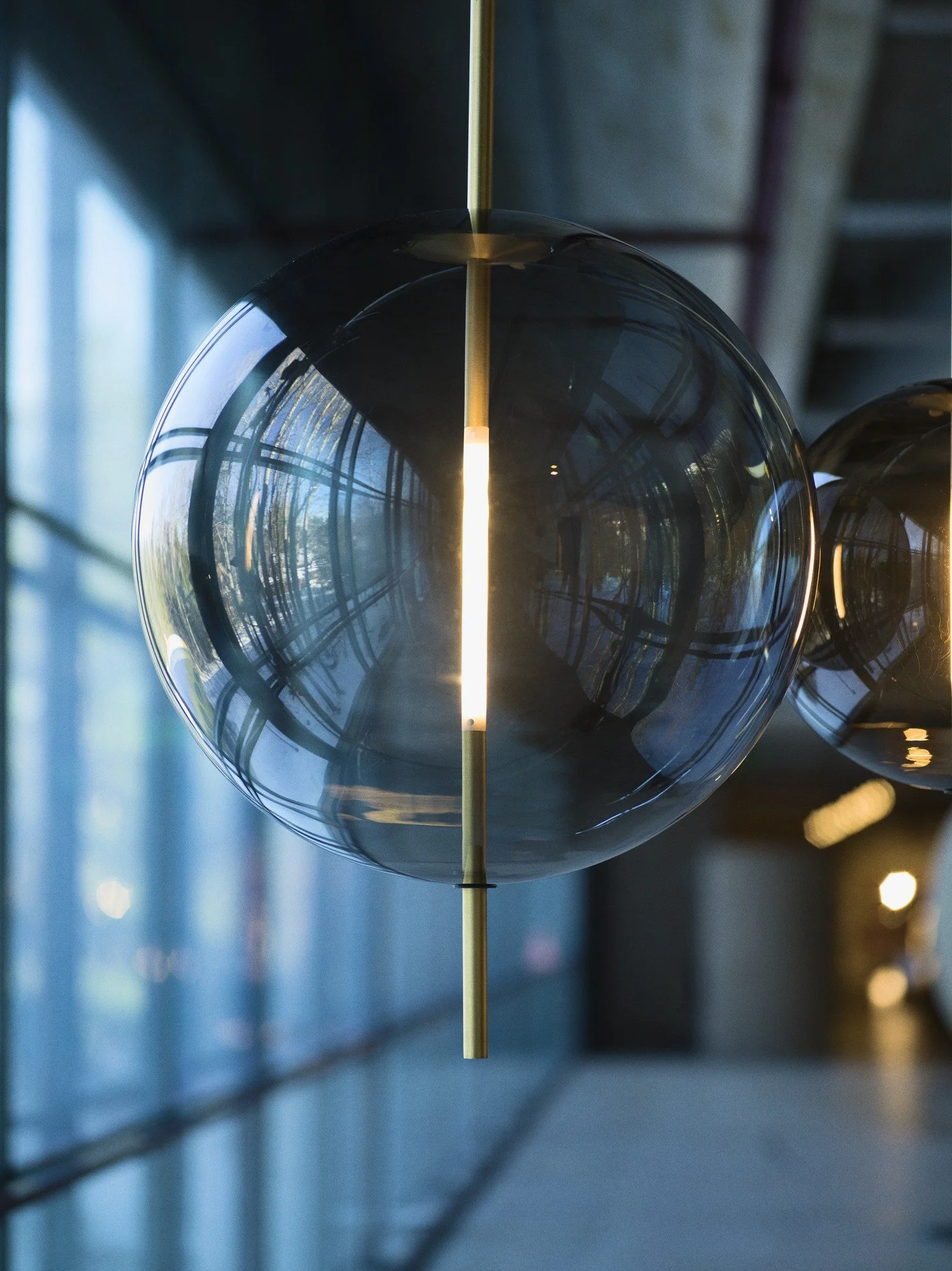 Close-up of two modern spherical hanging light fixtures with glass orbs and vertical gold rods, in an interior space with large windows and reflections.